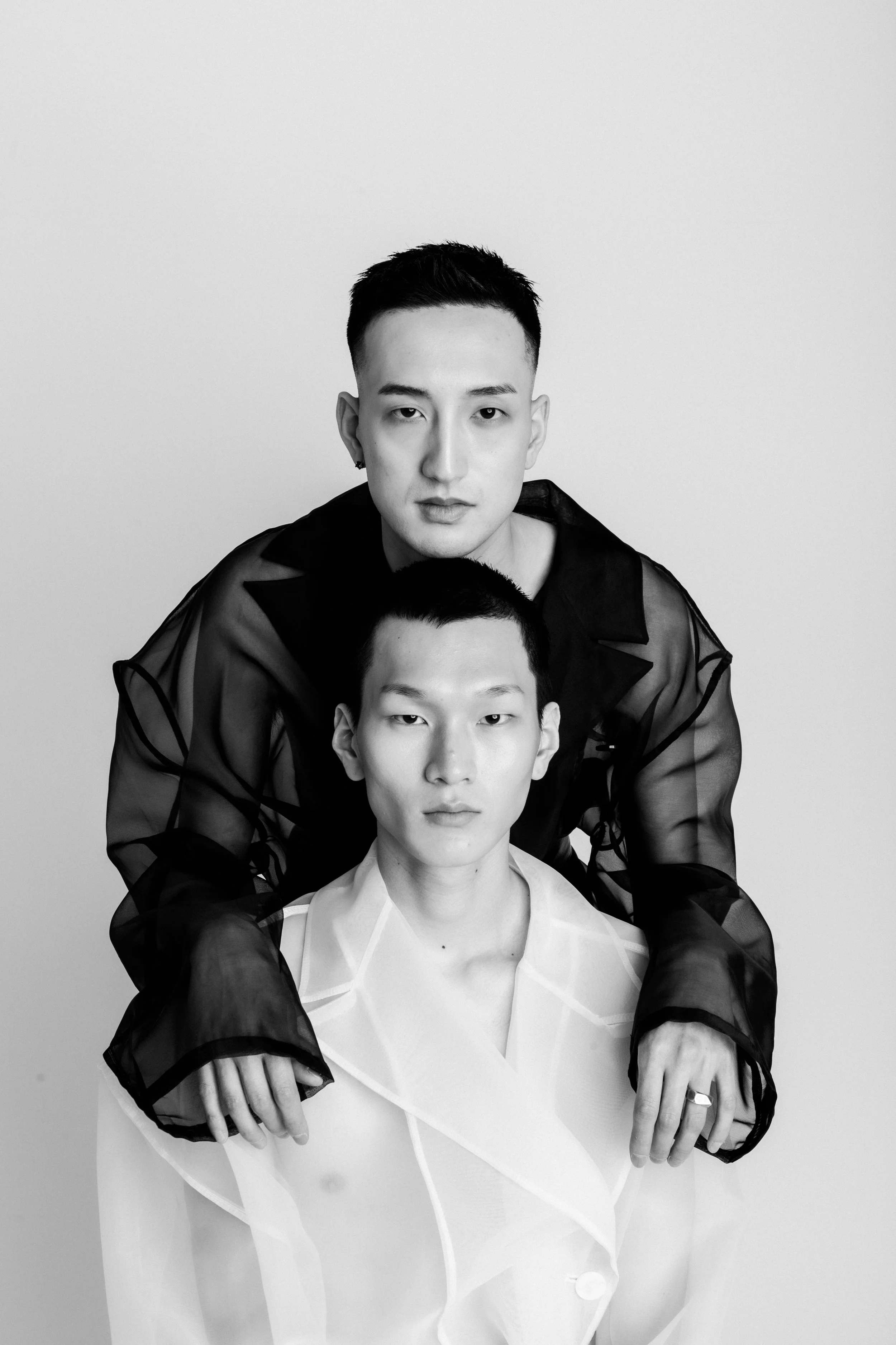 Two young men with short hair, one in a dark sheer top and the other in a light or transparent top, posing for a black-and-white portrait against a plain background.