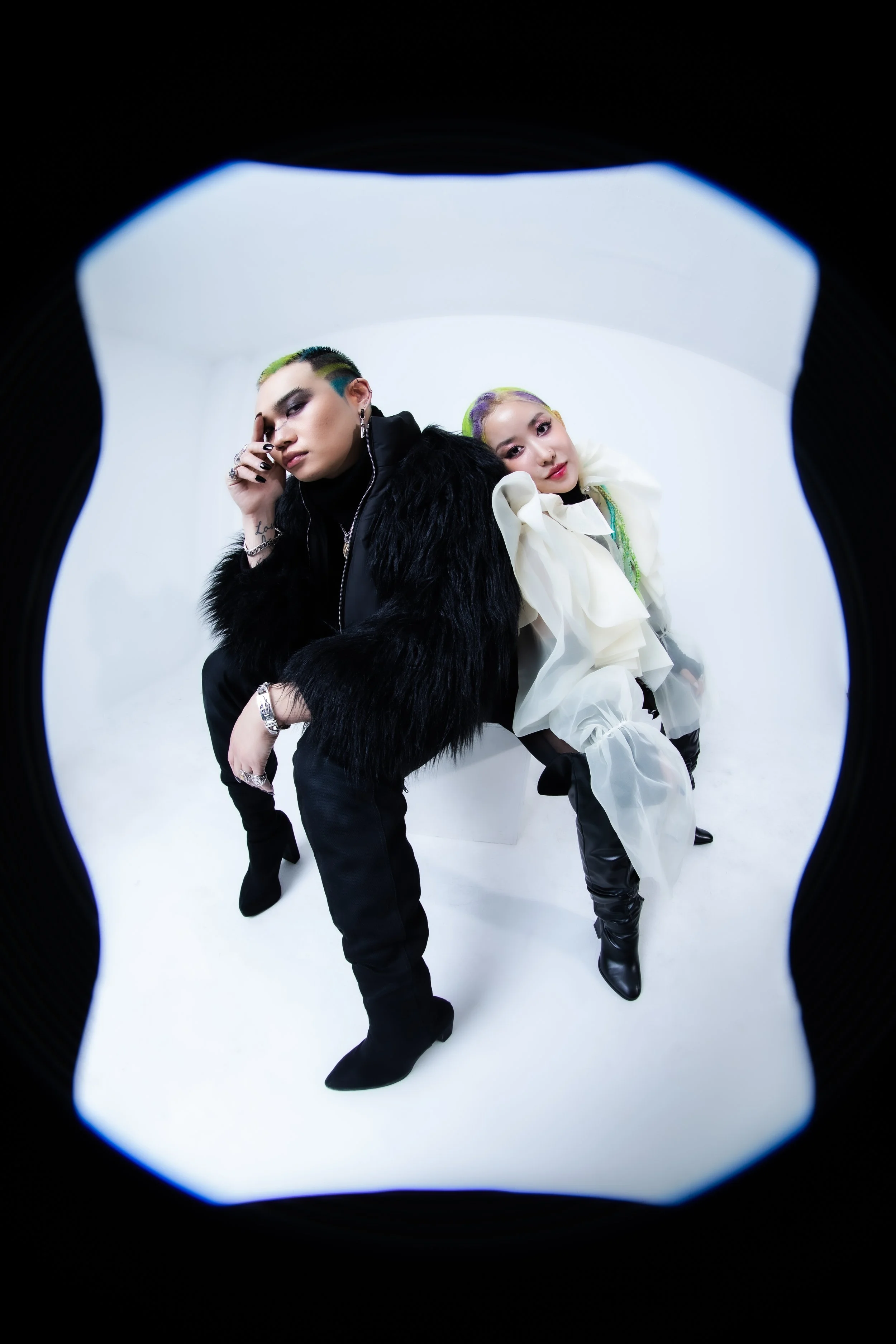 Two fashionable women sit back-to-back in a white photo studio, one with short green and black hair dressed in black fur coat, the other with long colorful hair in cream and black outfit.