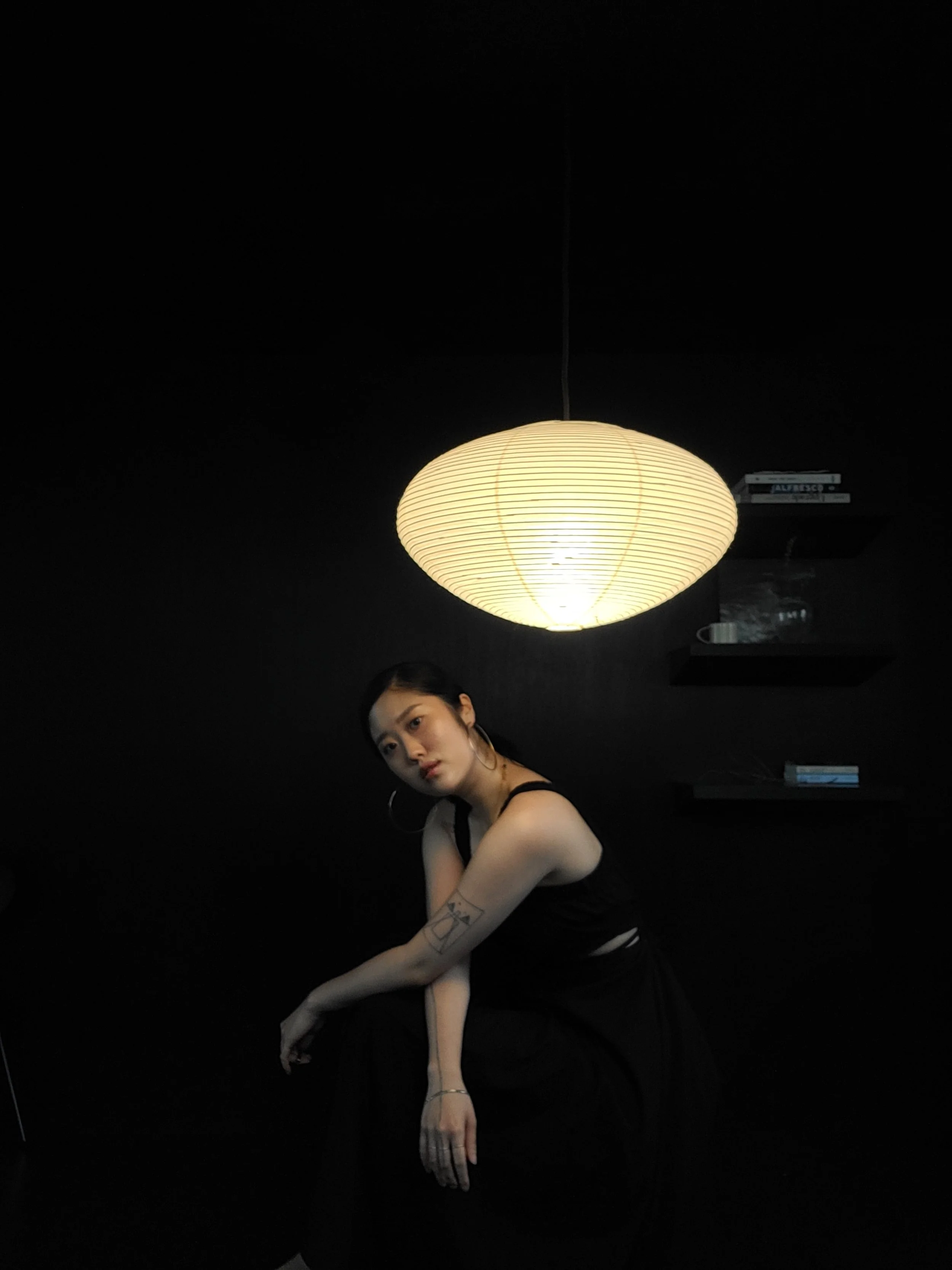 A young woman in black clothing and hoop earrings posing in front of a hanging paper lantern light against a dark background.