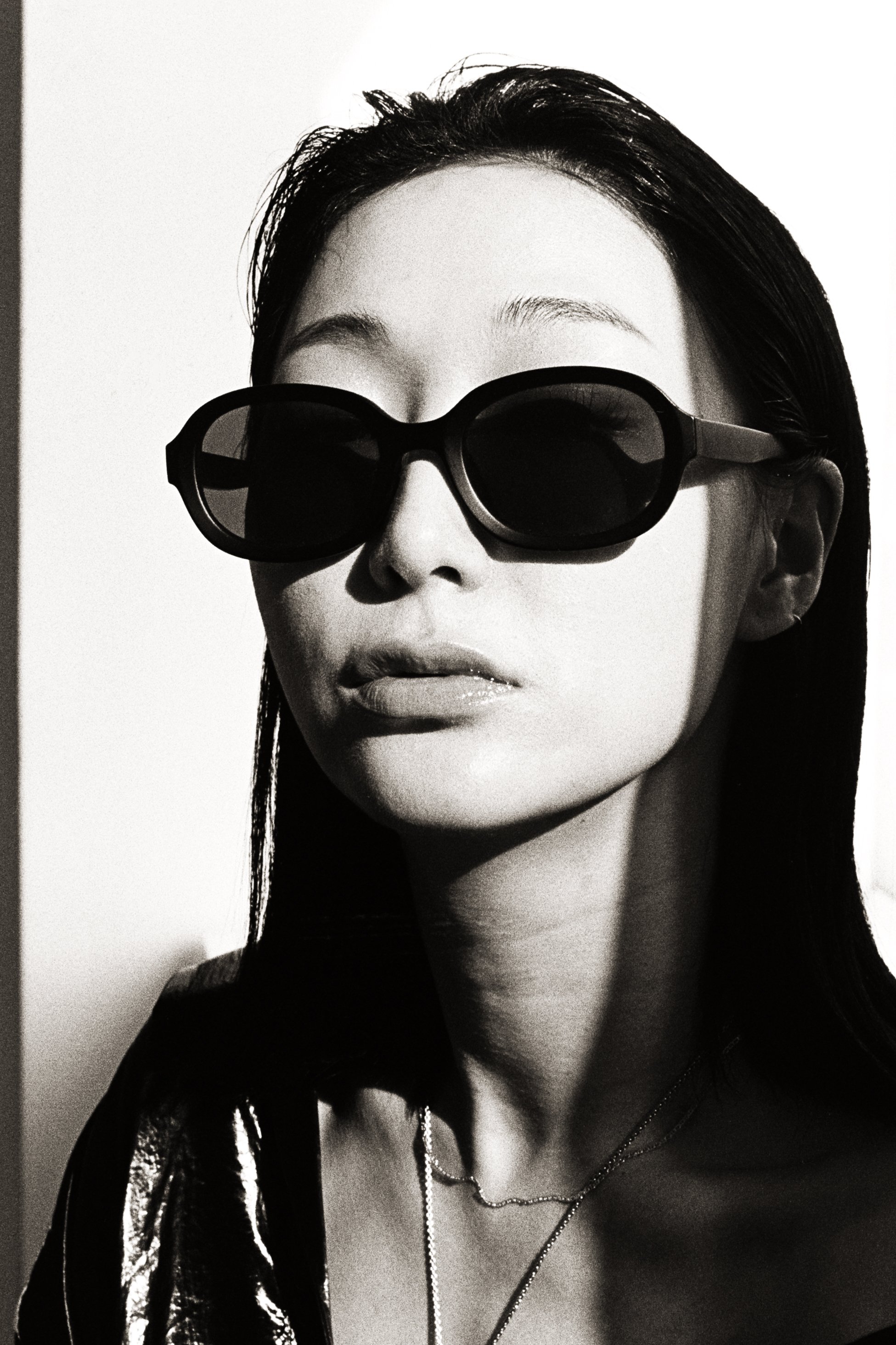 A black-and-white photo of a woman wearing large sunglasses, with sleek hair, lips slightly parted, and a shiny clothing top and necklaces.