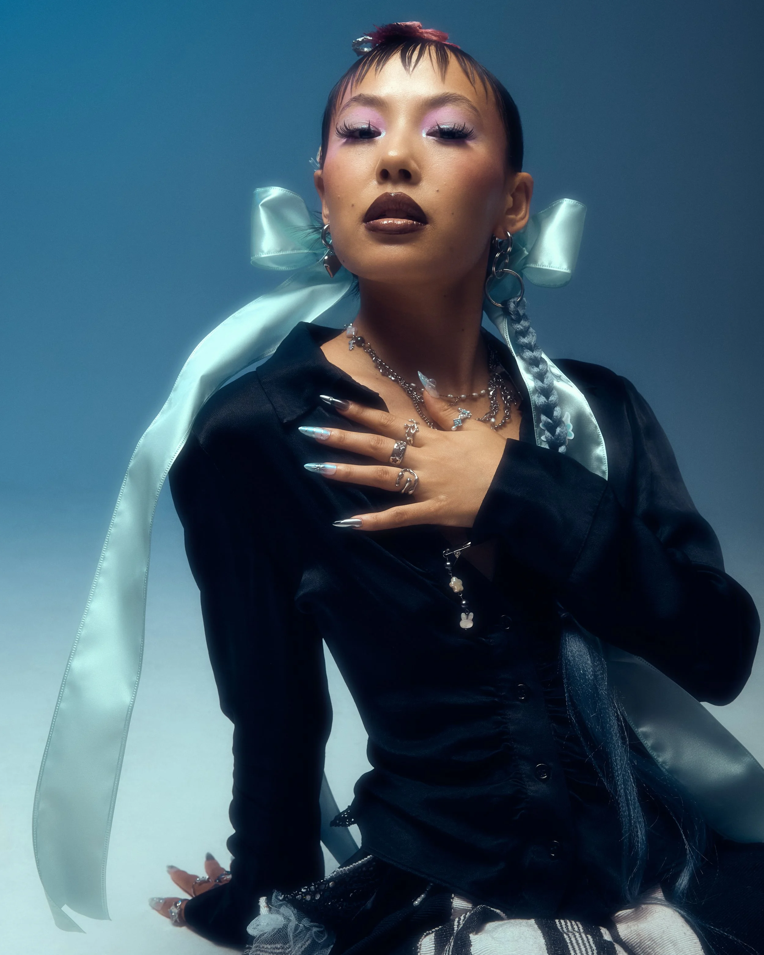 A woman with short hair styled with colorful braids and a satin ribbon, wearing dark lipstick, shiny eye makeup, and jewelry, striking a pose with one hand on her chest against a light blue background.