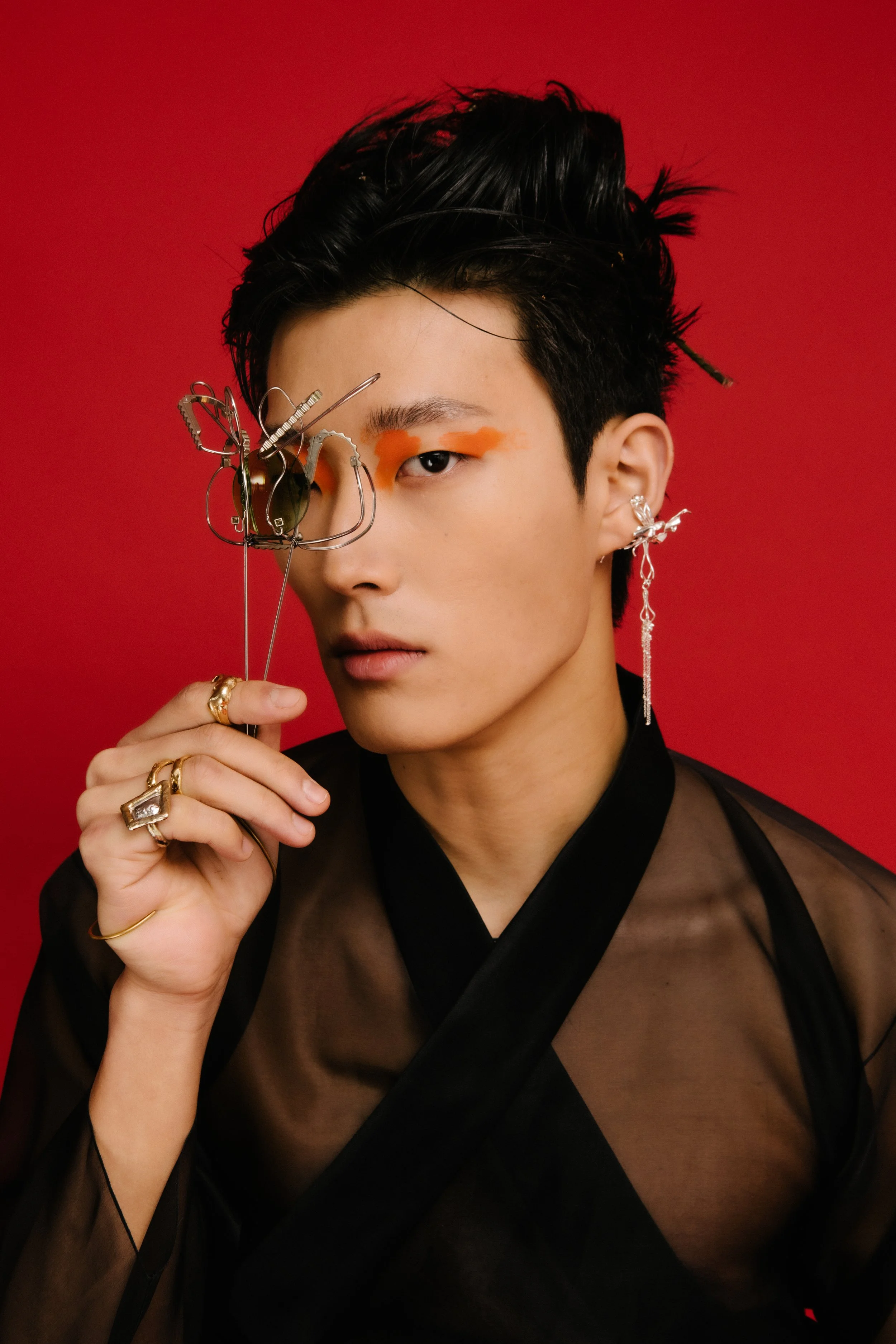 A person with short black hair, orange eye makeup, and multiple earrings holding a pair of glasses shaped like a butterfly in front of a red background.