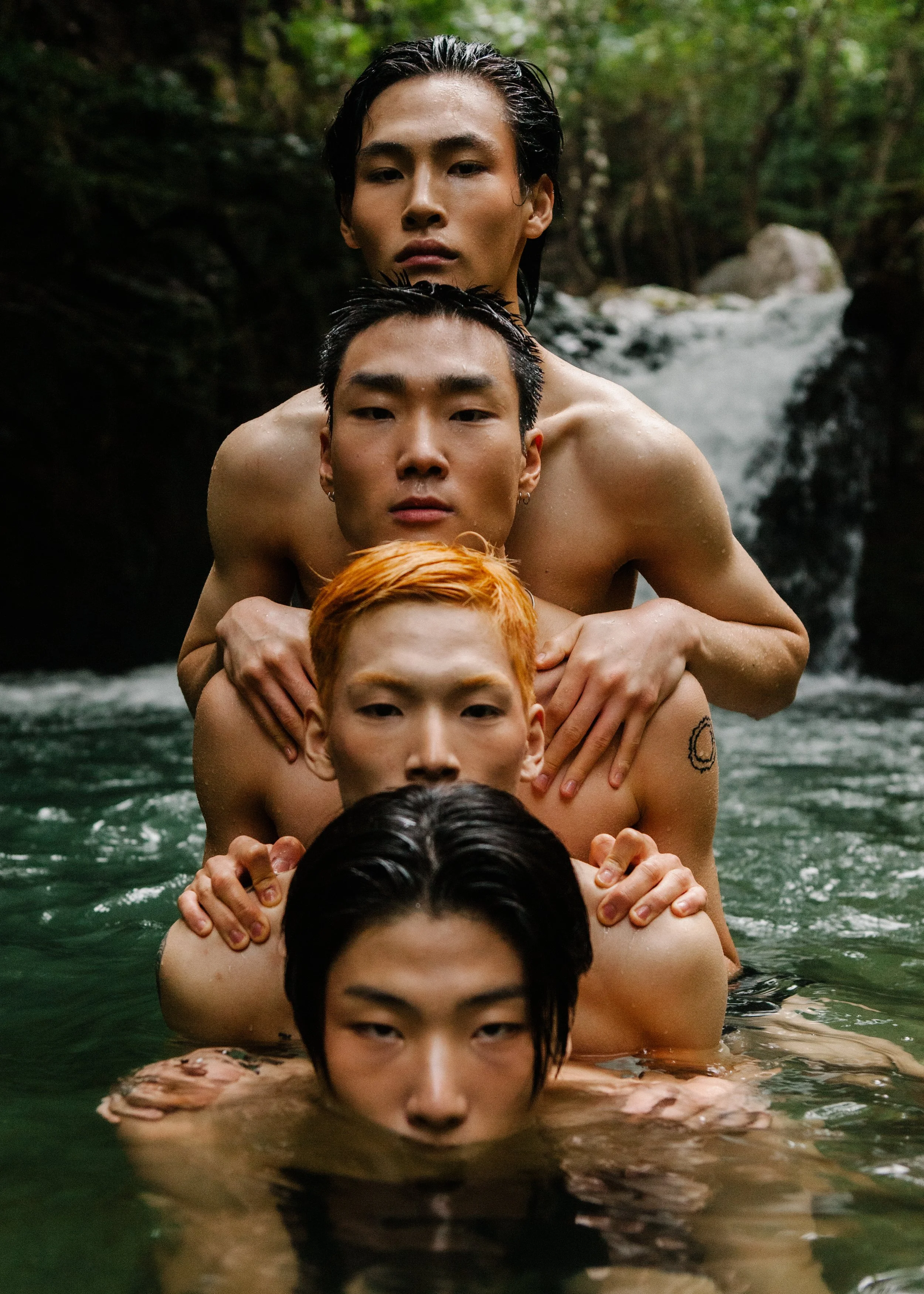 Five shirtless men with different hair colors and styles are standing in a river, one behind the other, with their hands on each other's shoulders. They are outdoors in a forested area with a small waterfall in the background.