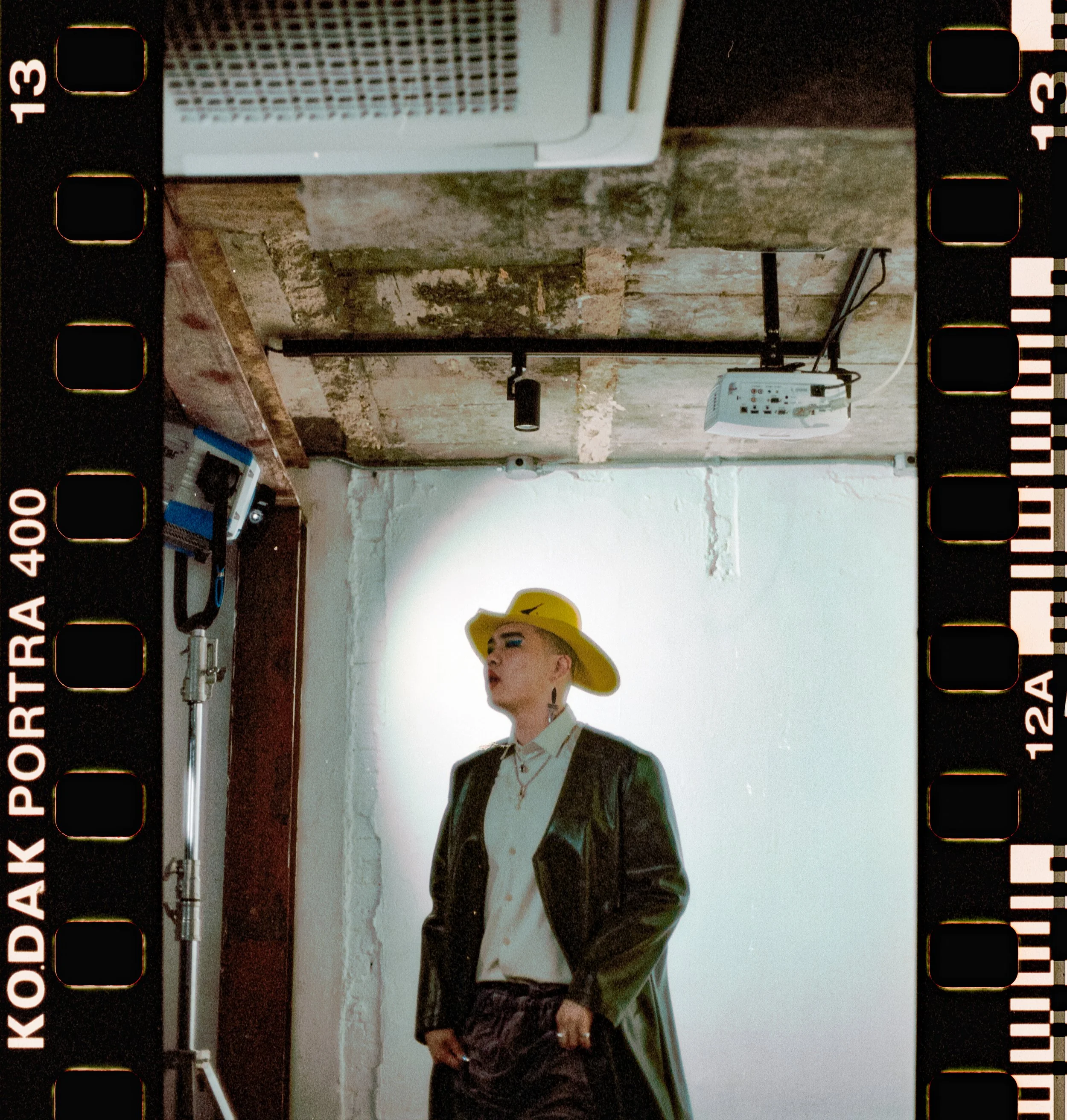 A person with short hair wearing a yellow wide-brimmed hat, white shirt, and black leather jacket. They are standing indoors against a white wall, with a projector and some audio equipment visible in the background.