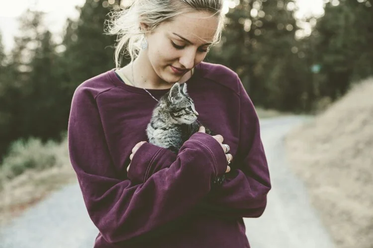 Young woman holding a small gray tabby kitten outdoors on a trail.