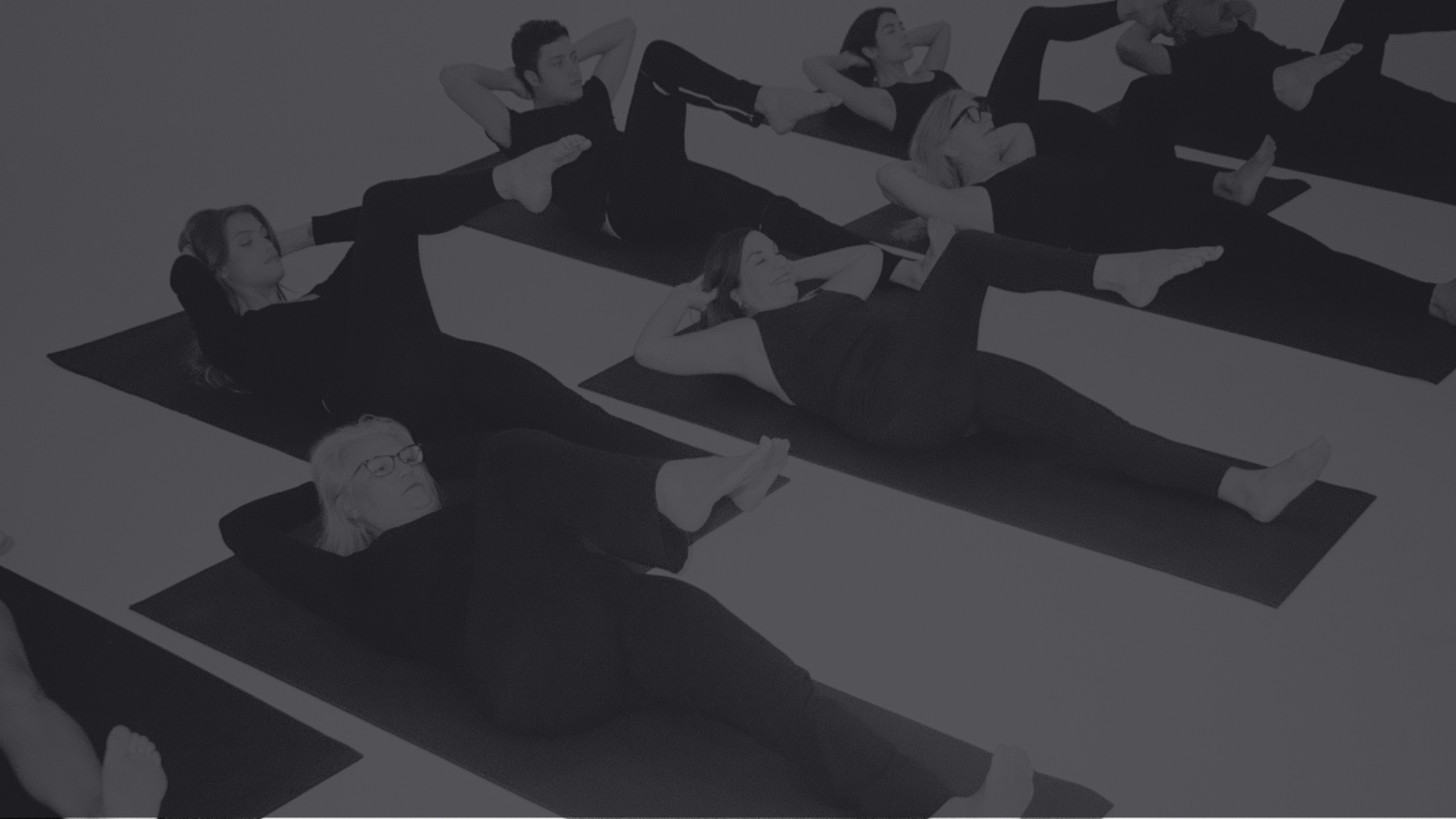 A group of women participating in a yoga or Pilates class, lying on black mats on the floor, performing leg and arm stretches.