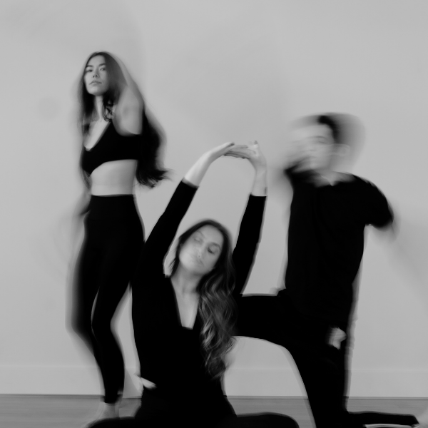 Three people in black clothing posing in a fitness studio, with one sitting on the floor and two standing, in a blurred black and white photograph.