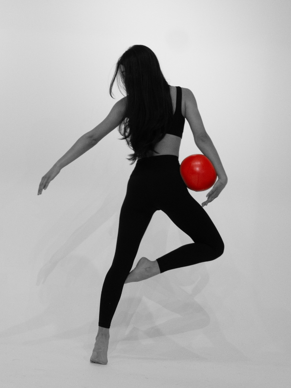 A woman in black athletic wear balancing on one foot, holding a red ball on her right side against a light background.