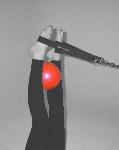 Person lying down with legs raised, holding a resistance band attached to a red exercise ball for fitness exercise.