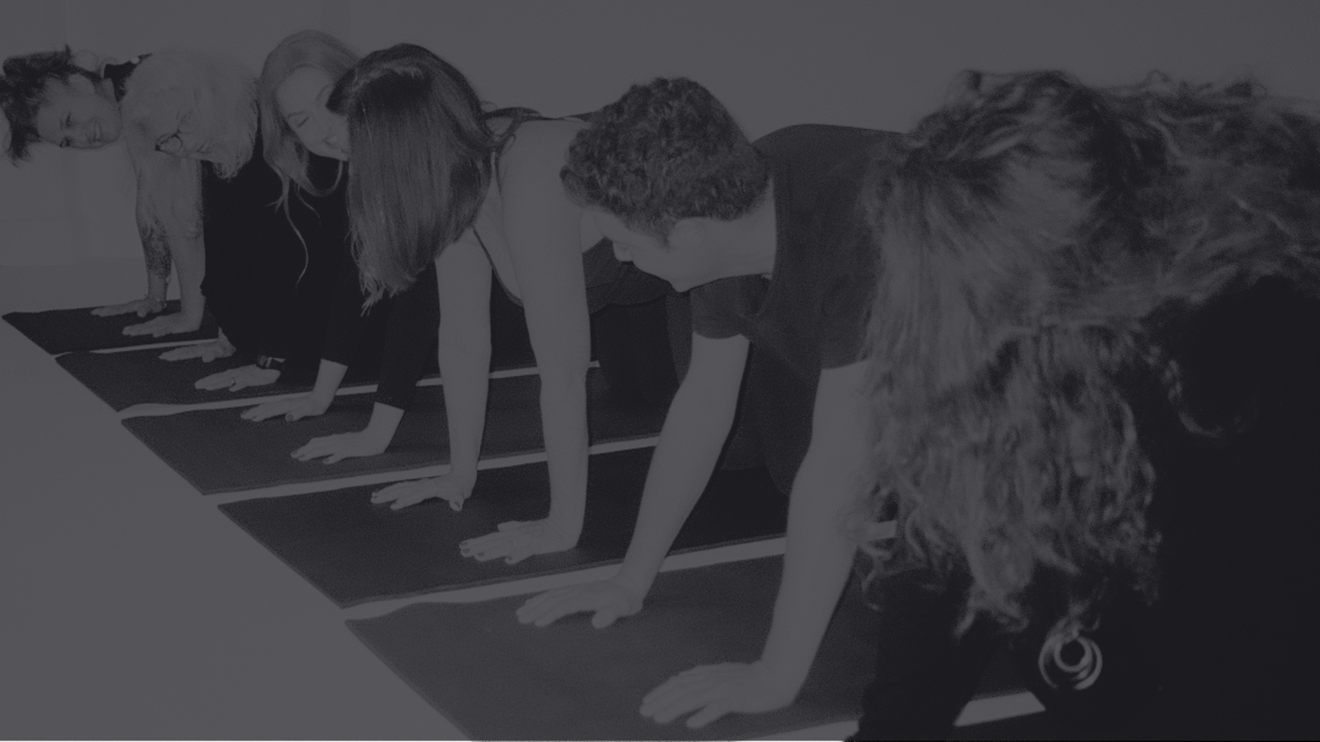 Group of six people practicing yoga on black mats, doing downward dog pose indoors.