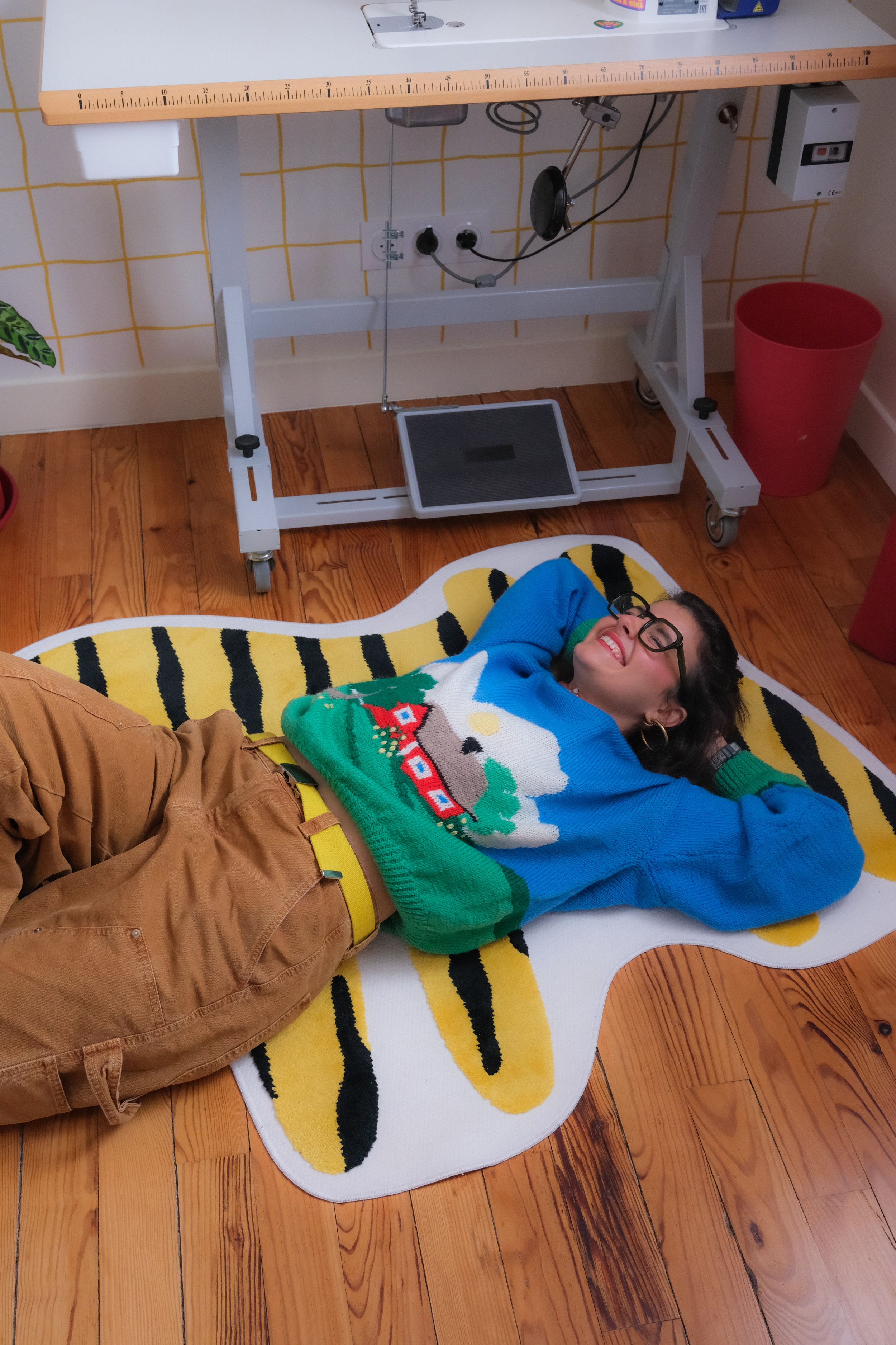 Une femme souriante, portant un pull coloré avec un motif d'oiseau, allongée sur un tapis en forme d'abeille, dans une pièce avec un sol en bois clair et un mobilier de style médical ou hospitalier.