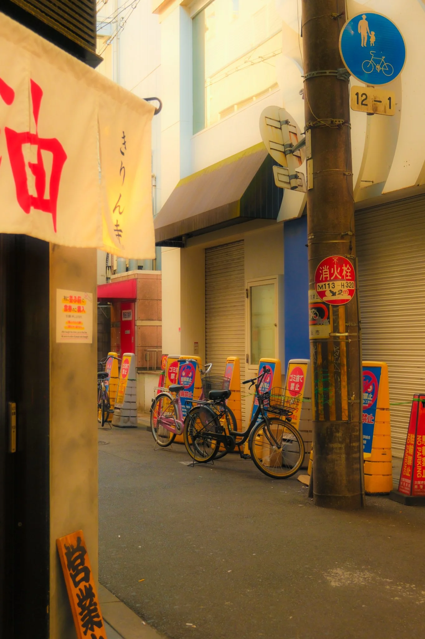 Une rue urbaine avec des vélos stationnés sur le trottoir, des panneaux de signalisation et un panneau en japonais, dans un environnement ensoleillé.