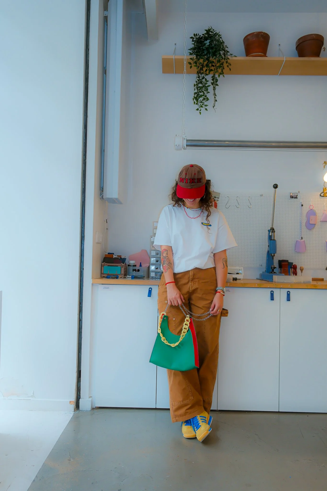 Jeune femme dans un atelier ou un studio créatif, portant un T-shirt blanc, un pantalon marron, un casque de baseball rouge, et tenant un sac à main coloré avec des chaînes.