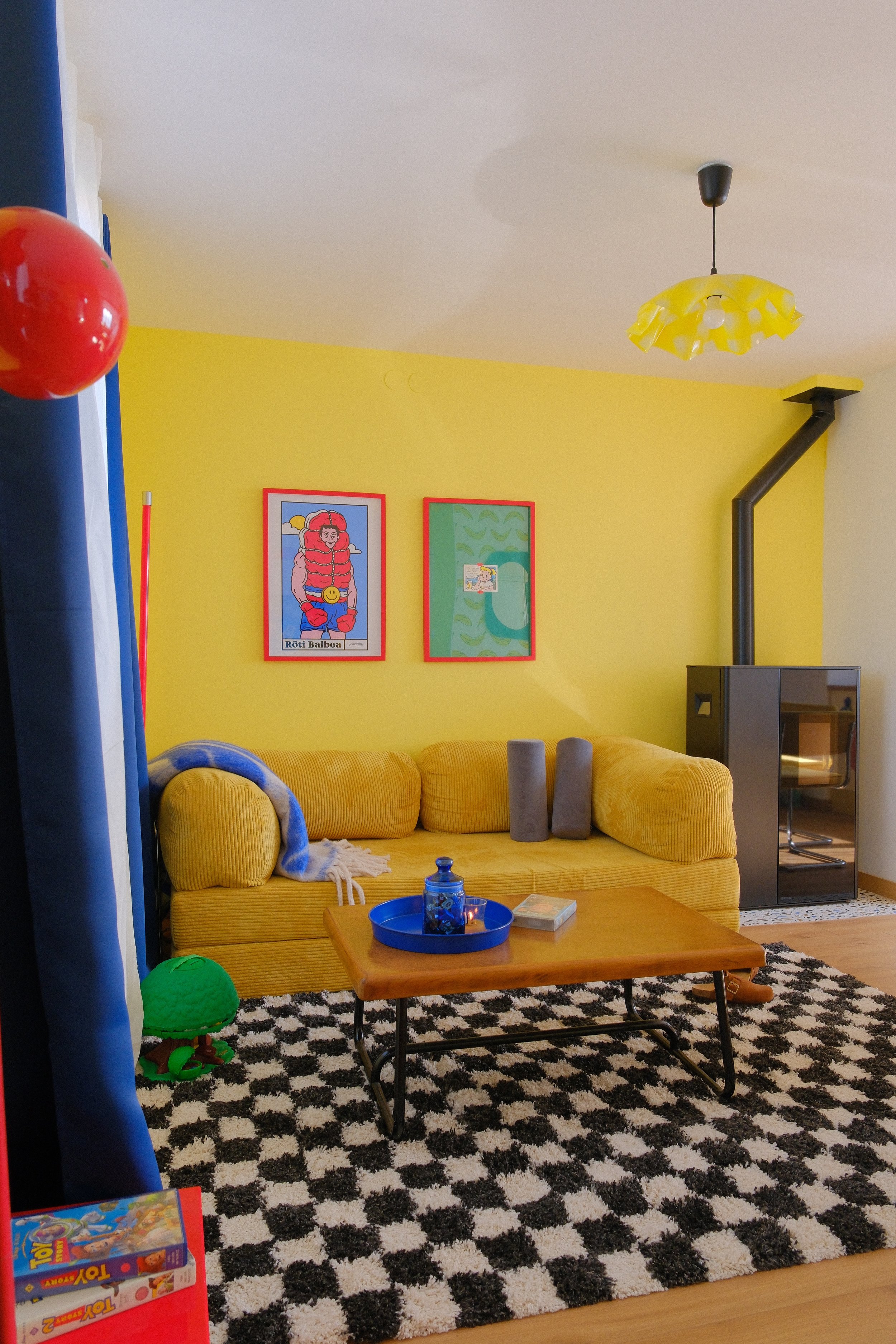 Un salon coloré avec un canapé jaune, un tapis noir et blanc, un tableau coloré sur le mur, une cheminée noire, et un plafonnier jaune.