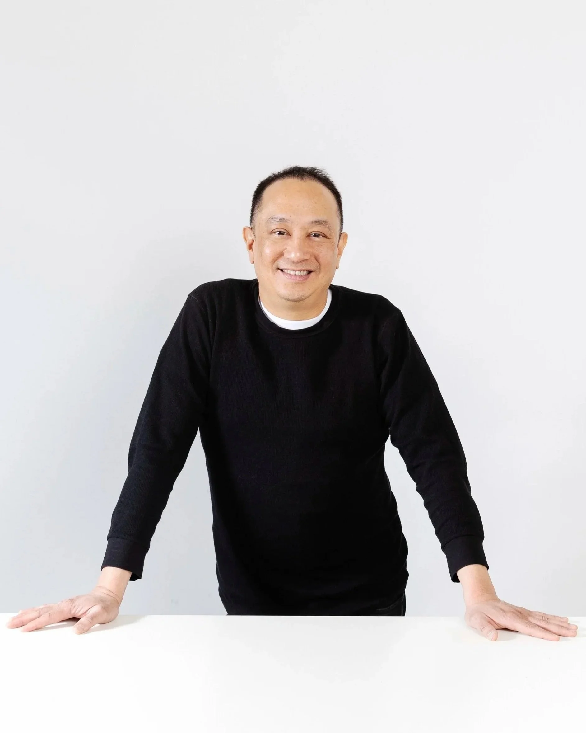 Un homme souriant, debout derrière une table blanche, appuyé sur ses mains.
