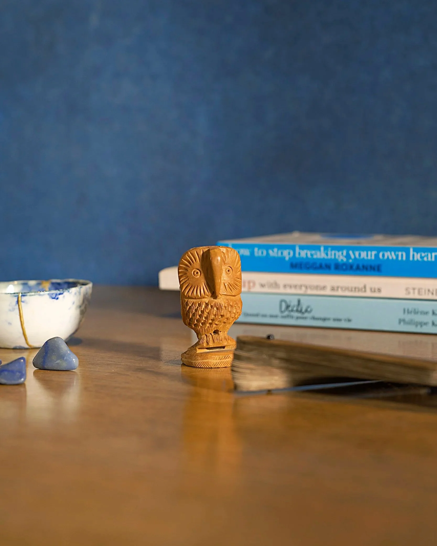 Une figurine en bois représentant une chouette, placée sur une surface en bois, avec un livre en arrière-plan et quelques objets en céramique bleue à gauche.