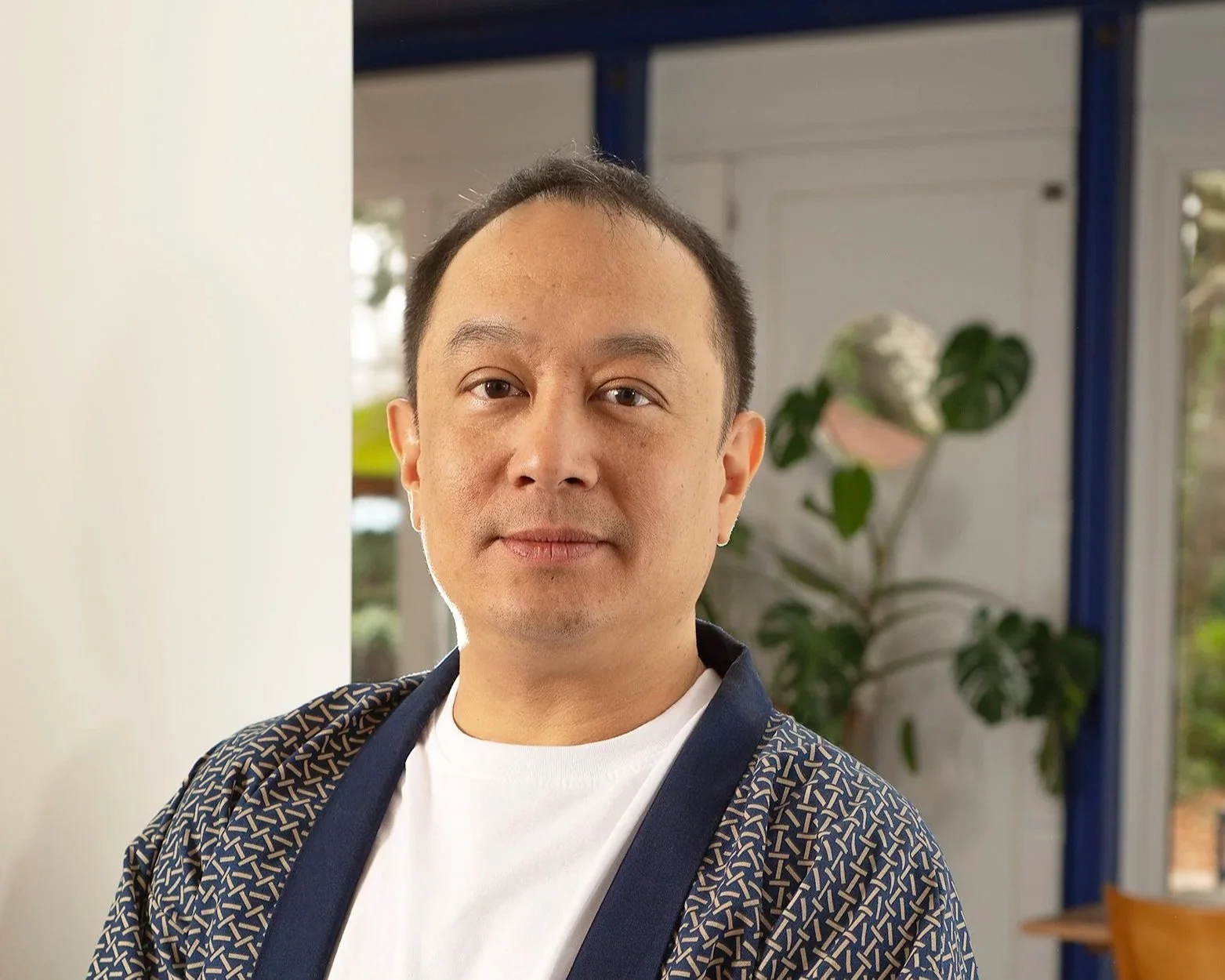 Un homme d'origine asiatique dans un intérieur moderne, portant une chemise à motifs et un t-shirt blanc, avec une plante verte en arrière-plan.