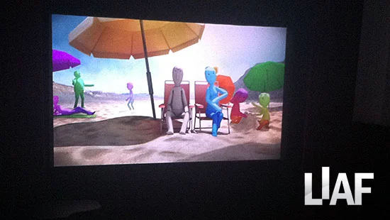 Scene from a computer animation featuring abstract, colourful characters from the short film 'A Little Grey' sitting on a beach with umbrellas.
