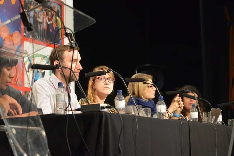 Panel of five people sitting at a long table with microphones, water bottles, and glasses, at a conference or discussion event.