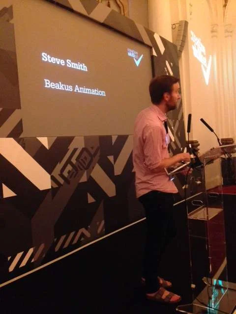 Beakus producer Steve Smith standing at a podium in front of a large screen displaying a presentation slide with the name 'Steve Smith' and the title 'Beakus Animation.'