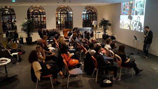 Group of people attending a presentation in a room with arched windows and potted plants, a speaker standing in front with a projected collage of images on the wall.