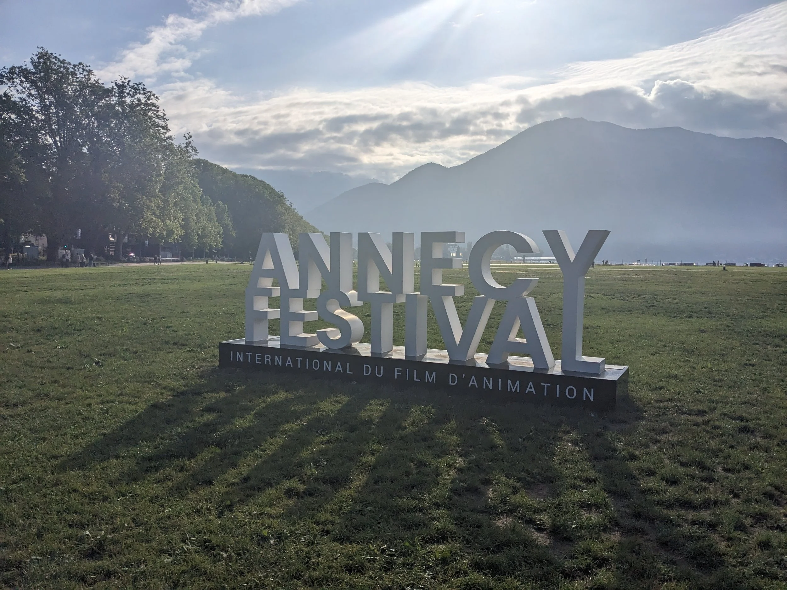 Sign for the Annecy International Animation Film Festival on a grassy area with mountains and a lake in the background.