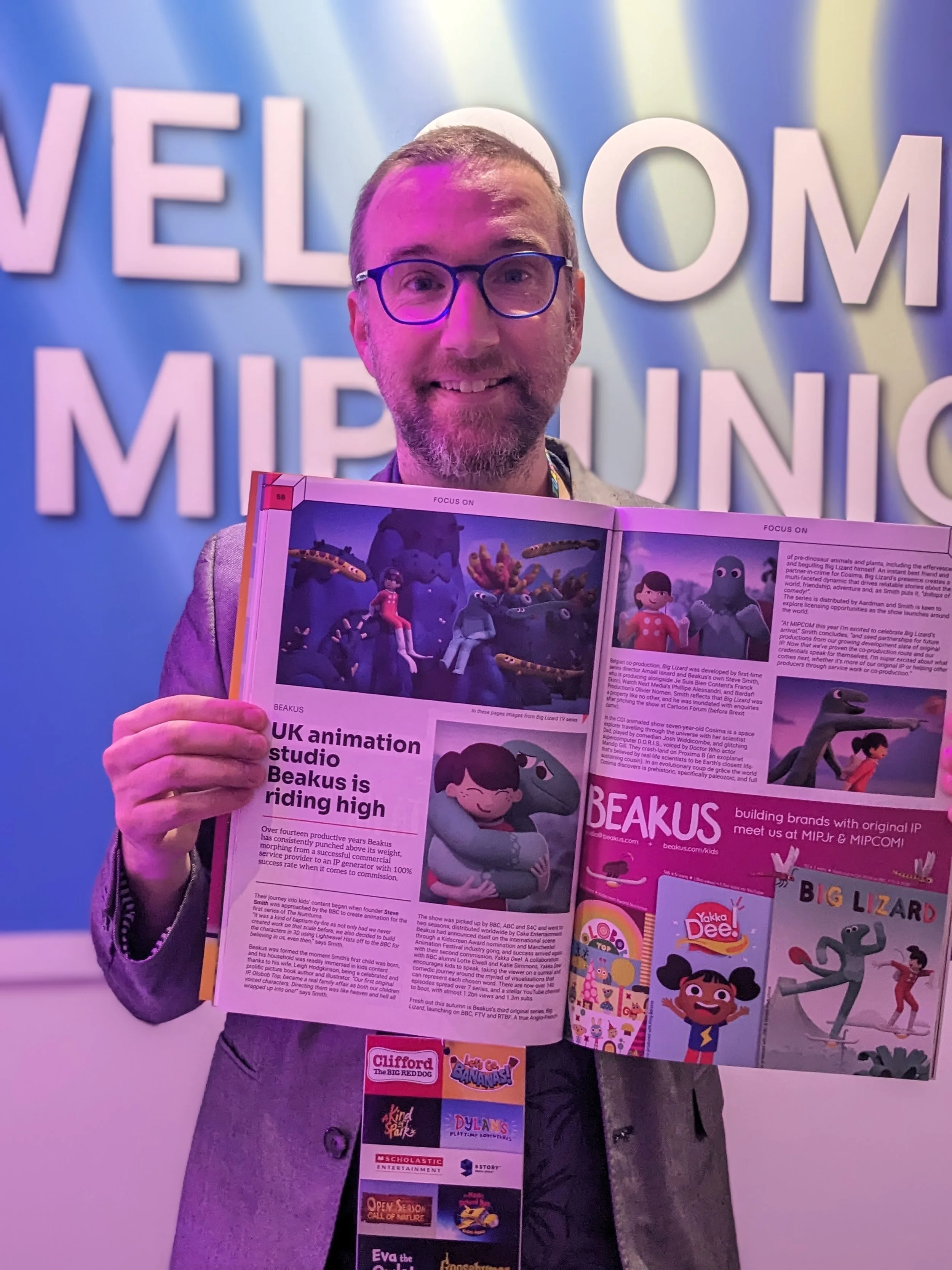 Steve Smith with glasses smiling, holding an open magazine with animated characters, standing in front of a colourful 'Welcome to MIPJR' sign.