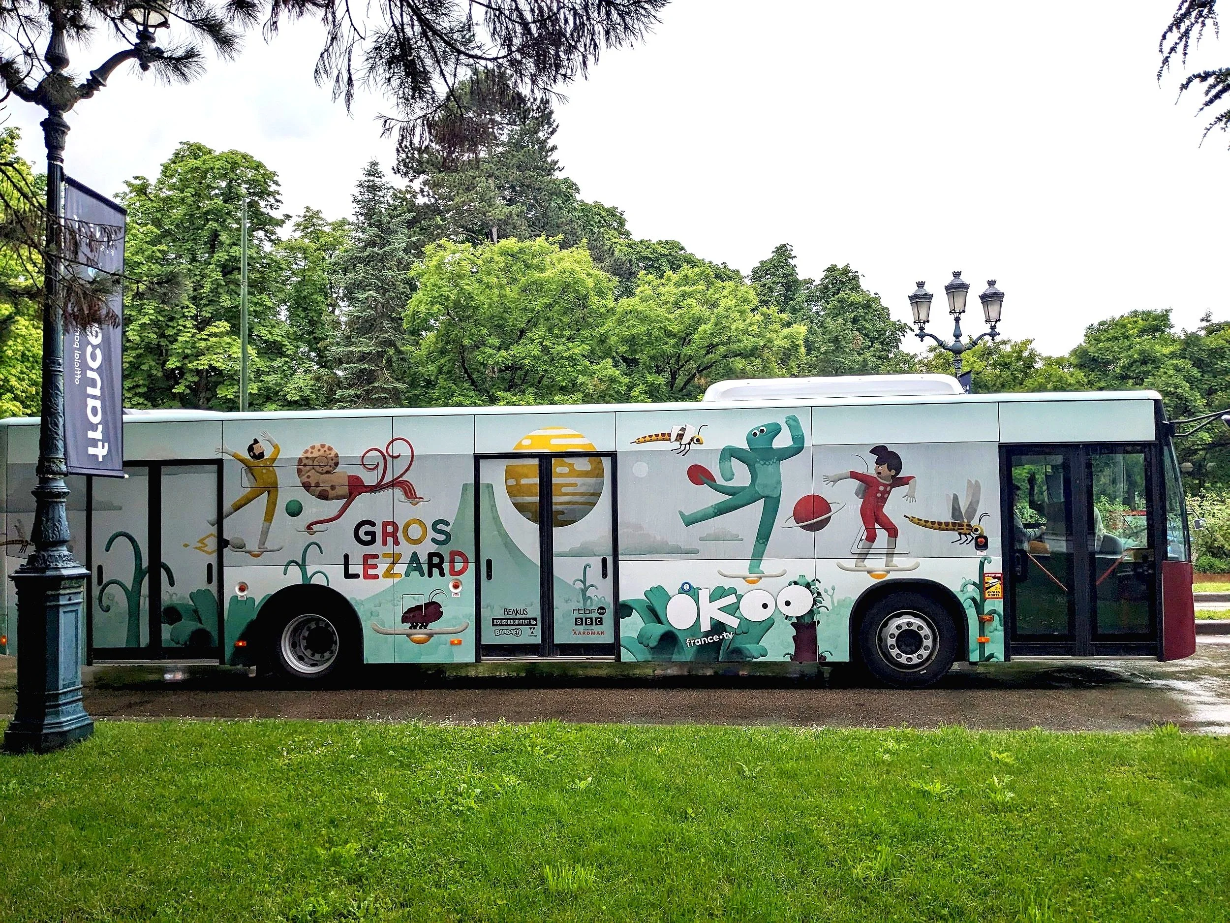 A colourful bus with illustrations from Big Lizard, parked on a street in a park, with green trees and grass around and a street lamp nearby.