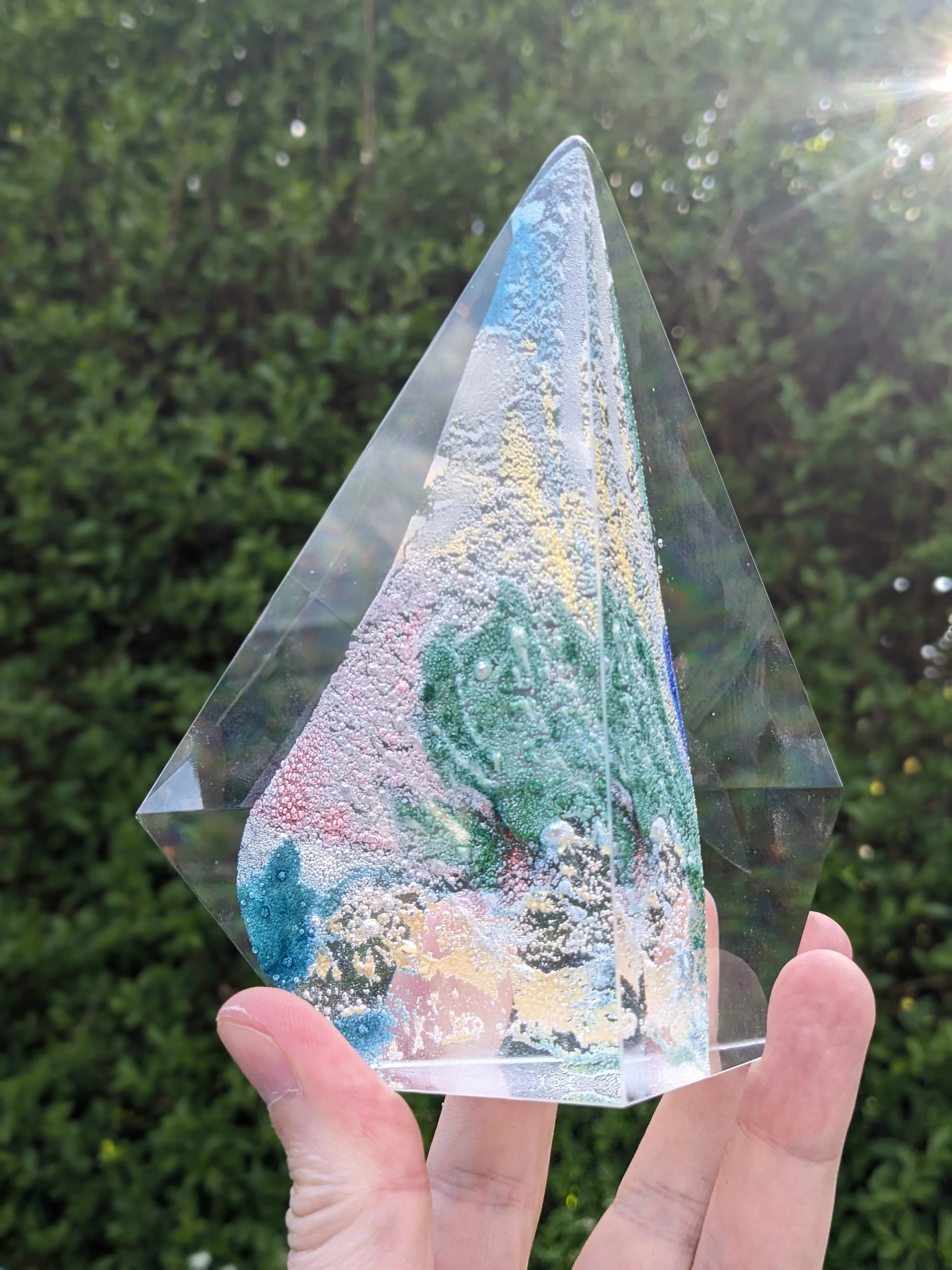 An Annecy International Animation Festival crystal award: a clear, transparent pyramid-shaped object held in a person's hand outdoors with green foliage in the background.