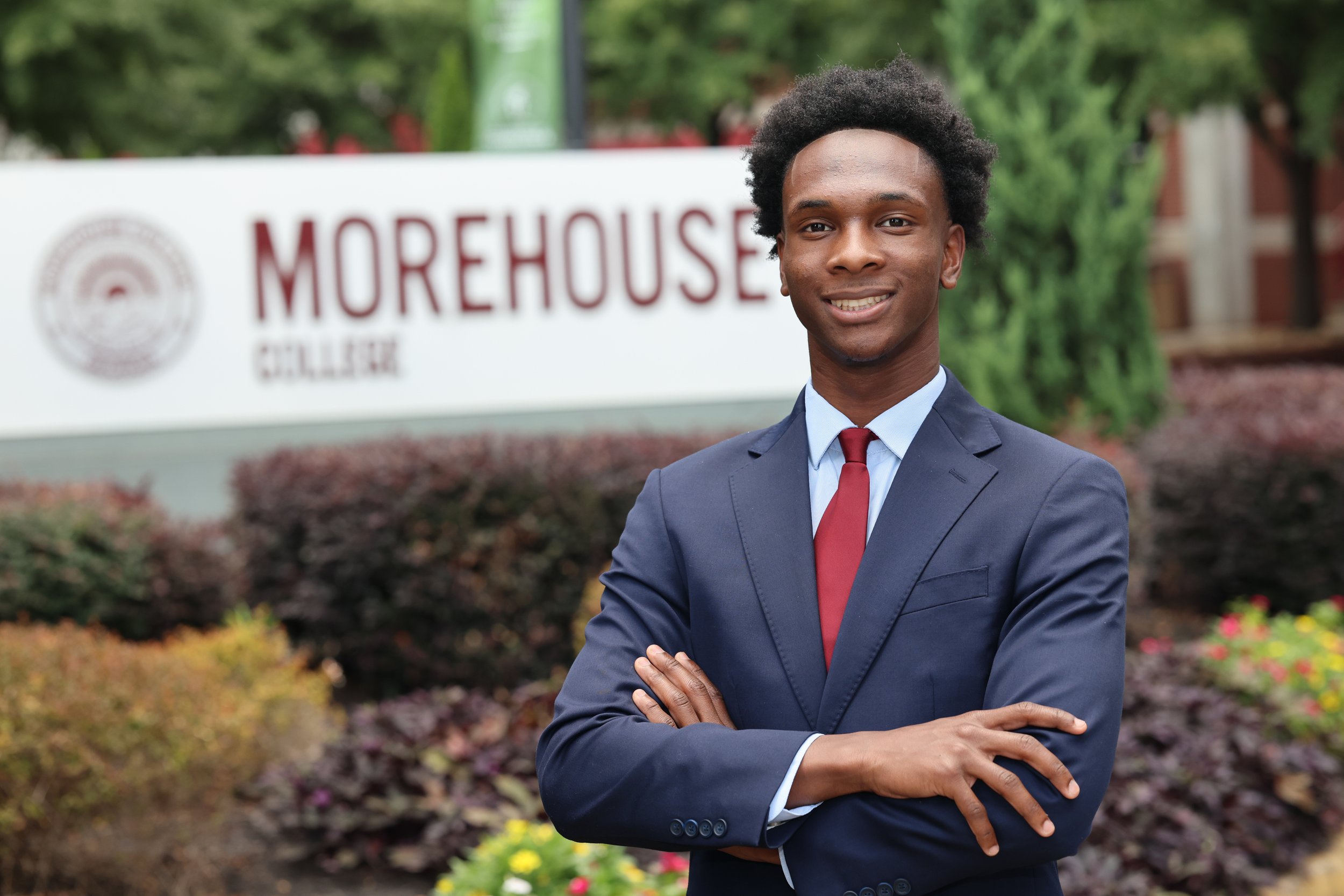 A young man in a navy blue suit and red tie standing outdoors with arms crossed, in front of a sign for Morehouse College with greenery in the background.