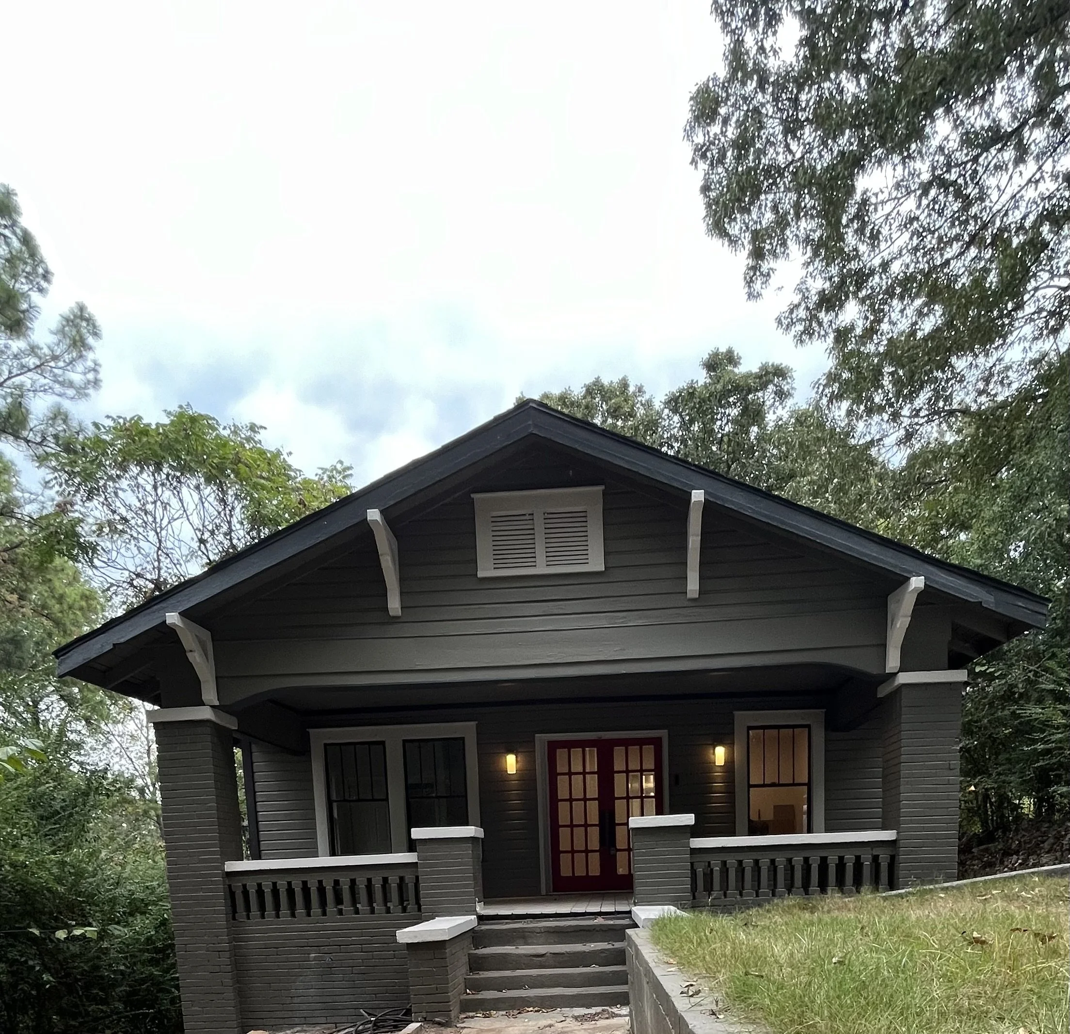 Front view of a dark gray house with a porch, steps, red front door with glass panels, and surrounding trees.