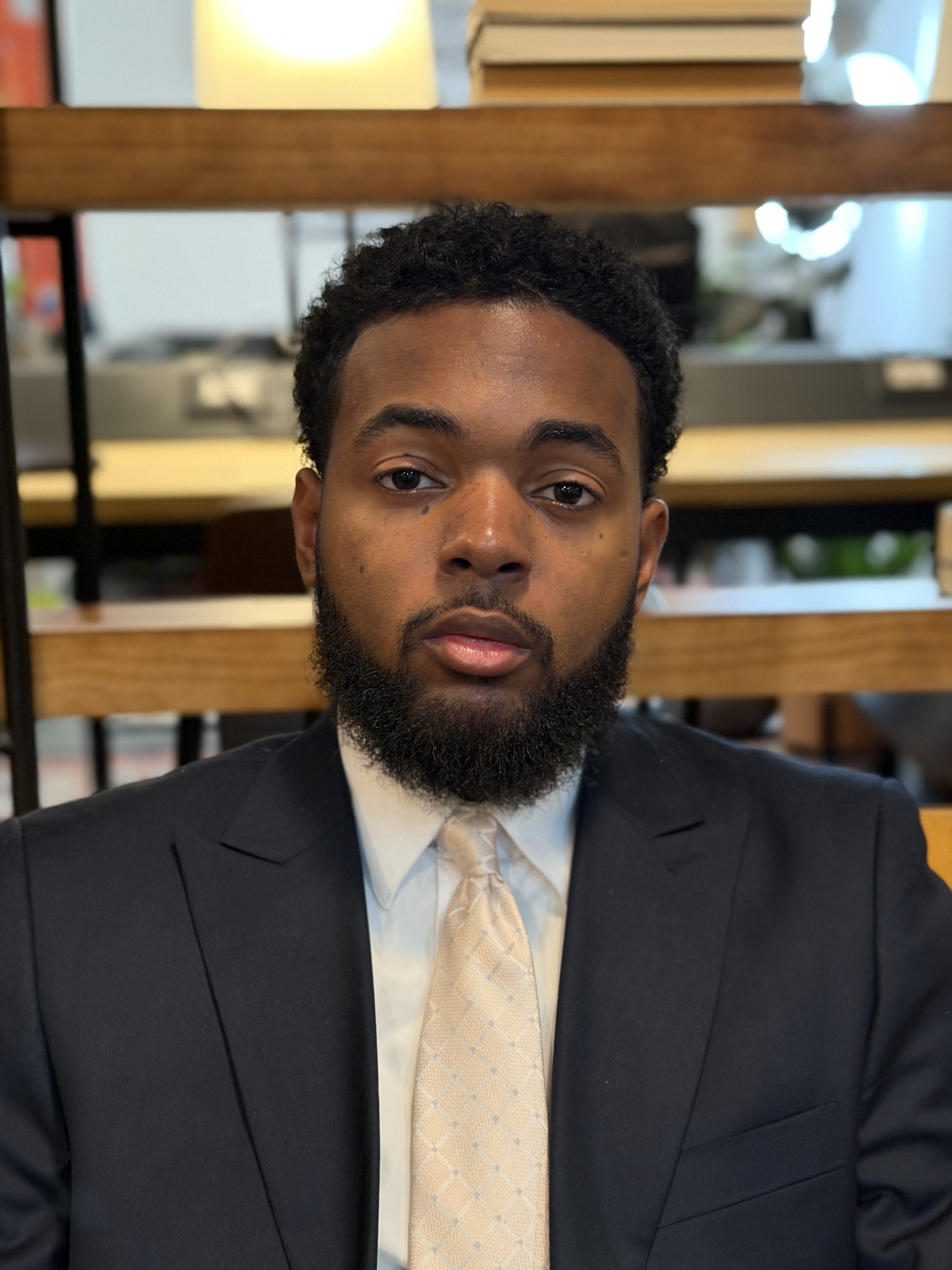 A man with a beard and short curly hair, wearing a black suit, white shirt, and beige tie, sitting indoors with wooden shelves and blurred background behind him.