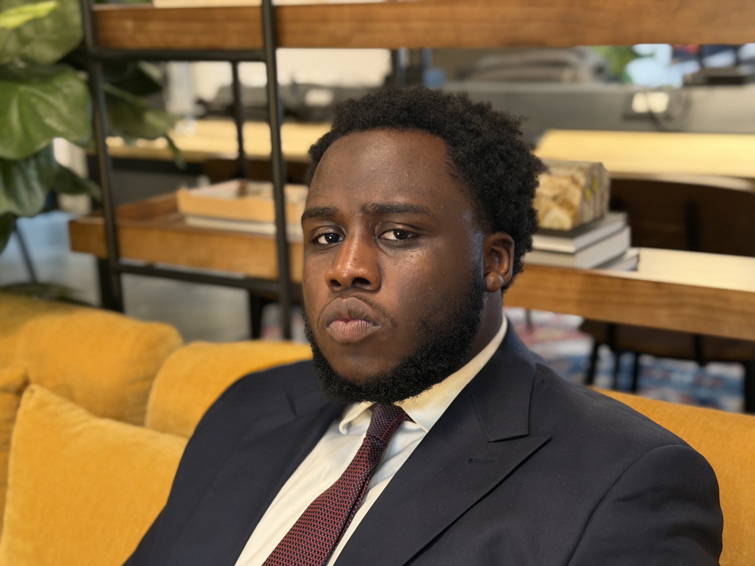 A young man with dark skin, black curly hair, and a beard, wearing a black suit, white shirt, and red tie, is sitting on an orange couch in a modern indoor space with wooden shelves and plants in the background.