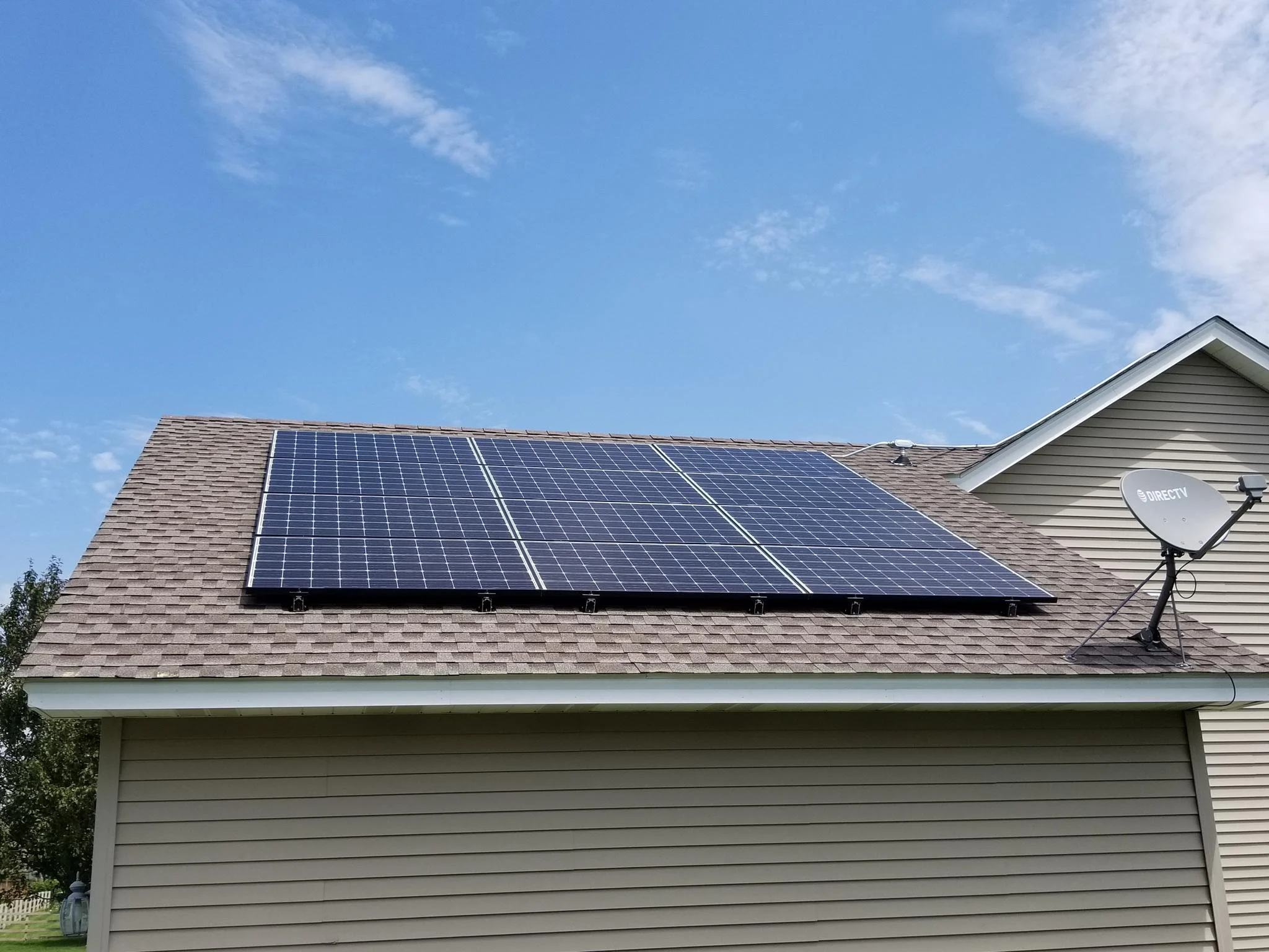 House with solar panels on the roof and a DirecTV satellite dish attached, under a blue sky with some clouds.