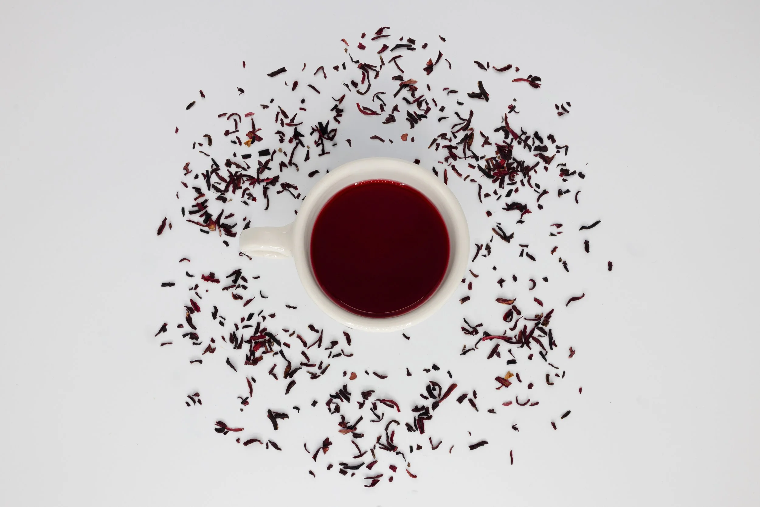 A white cup filled with red herbal tea surrounded by scattered dried hibiscus petals on a white surface.