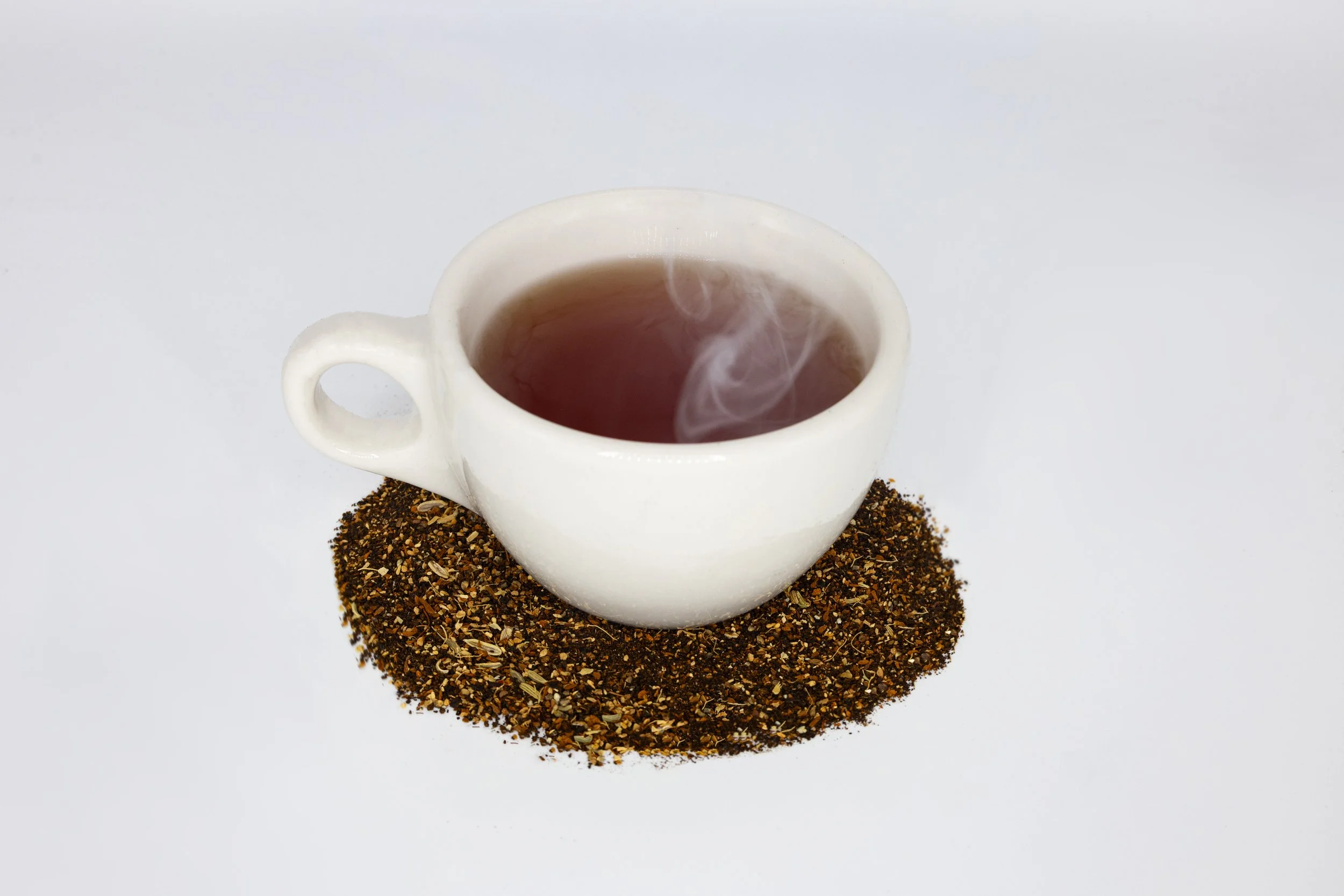 White ceramic cup filled with hot tea sits on a bed of loose tea leaves and spices on a white background.