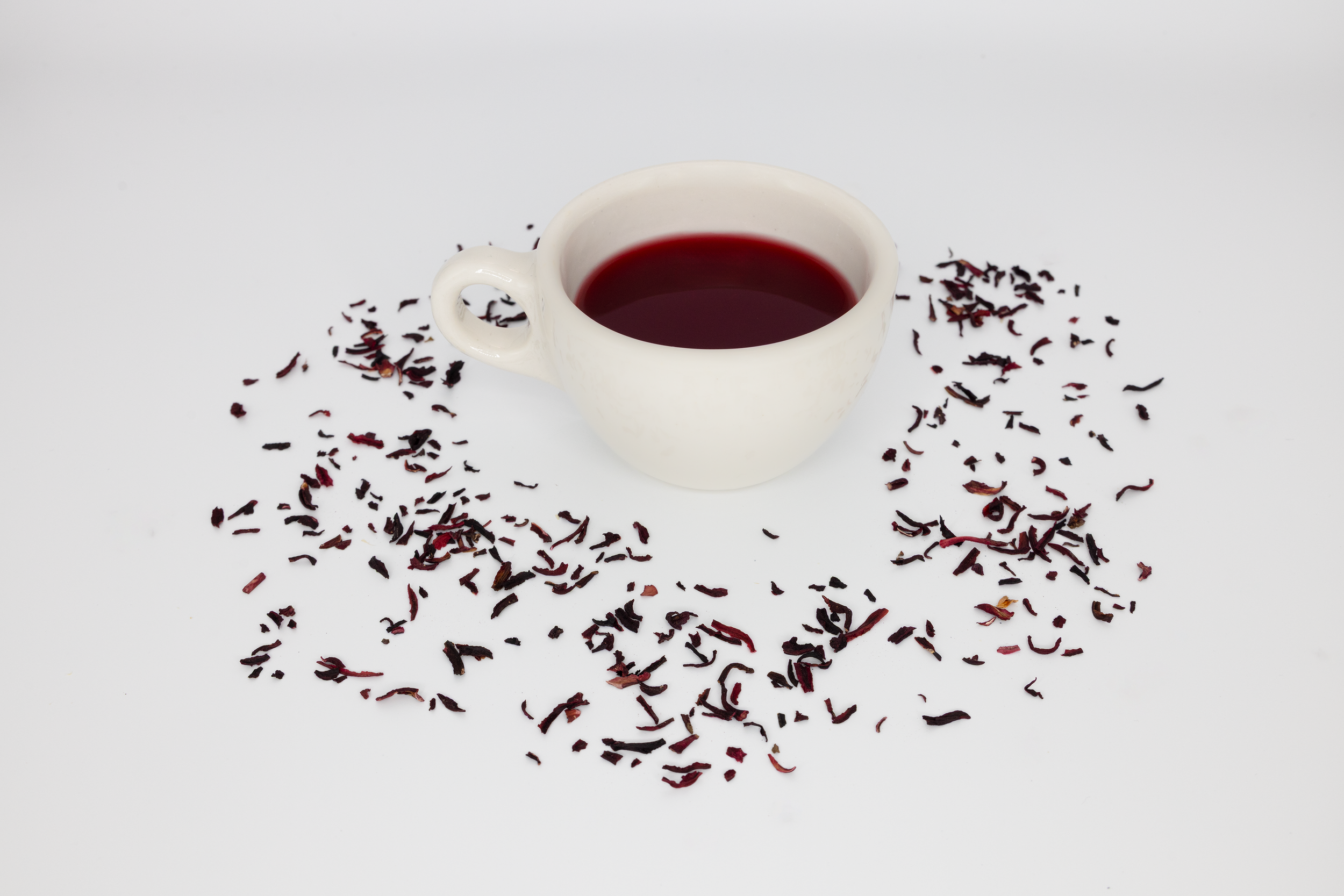 A white cup filled with dark red liquid, surrounded by scattered dried flower petals on a white background.