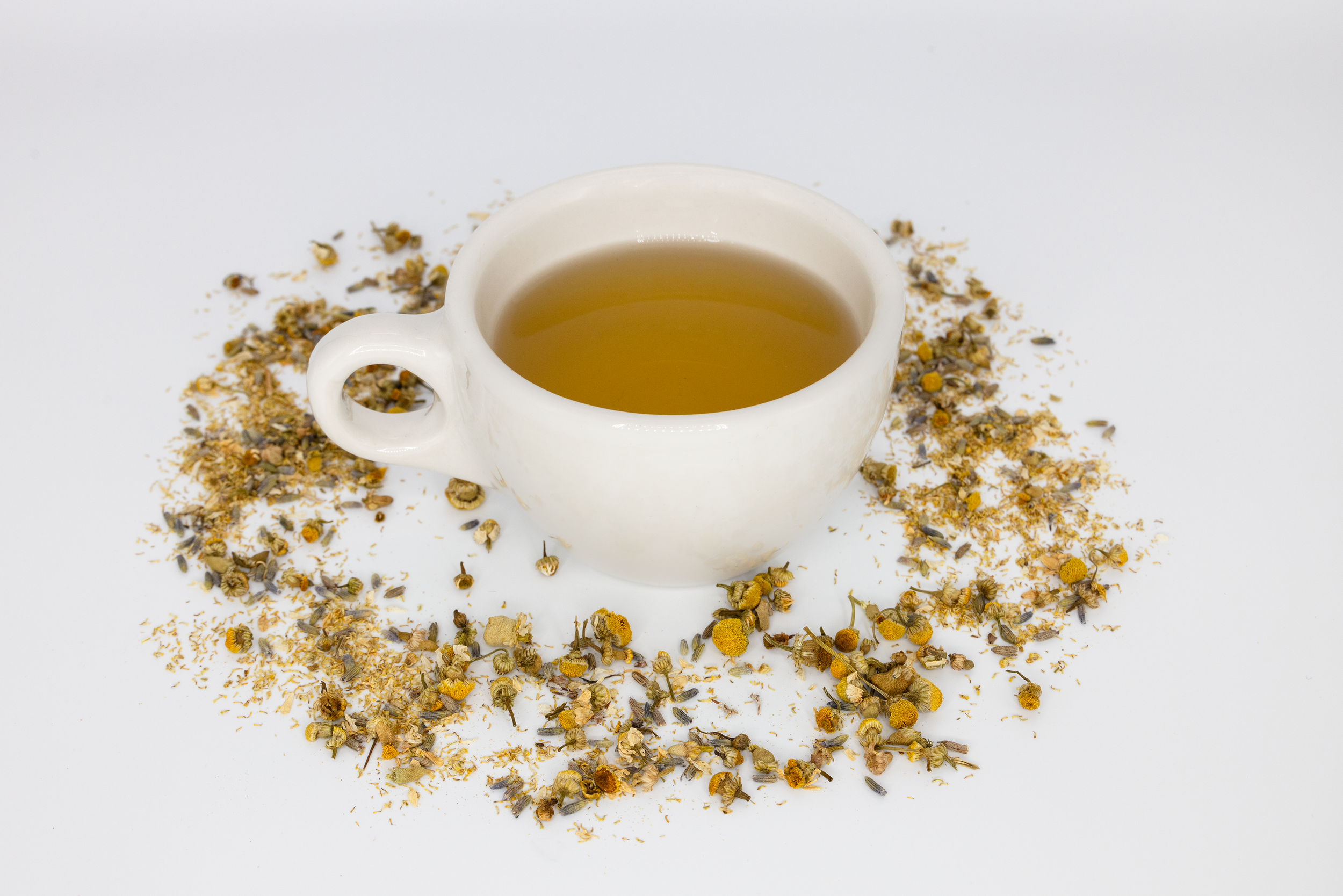 A white ceramic cup filled with herbal tea, surrounded by dried chamomile and floral herbs on a white background.
