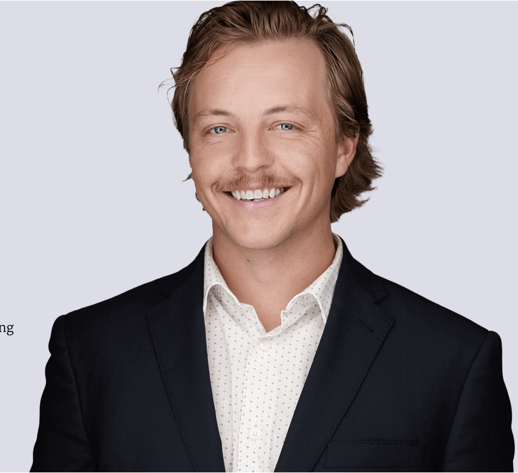A smiling man with light skin, light brown hair, and a mustache, wearing a dark blazer over a light patterned shirt.