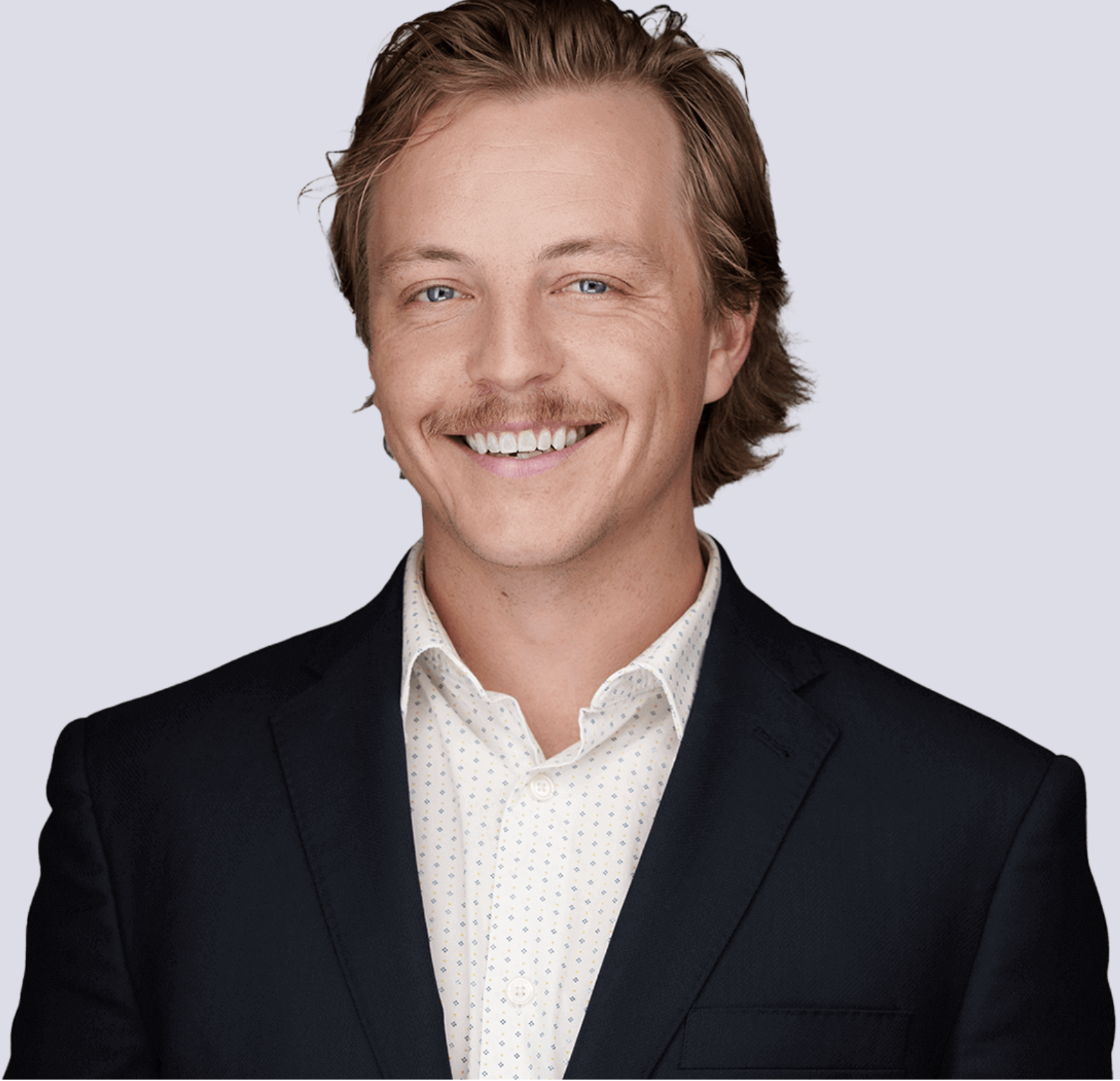 Headshot of a smiling young man with light brown hair, blue eyes, and a mustache, wearing a dark blazer and a white shirt with small pattern, against a light background.