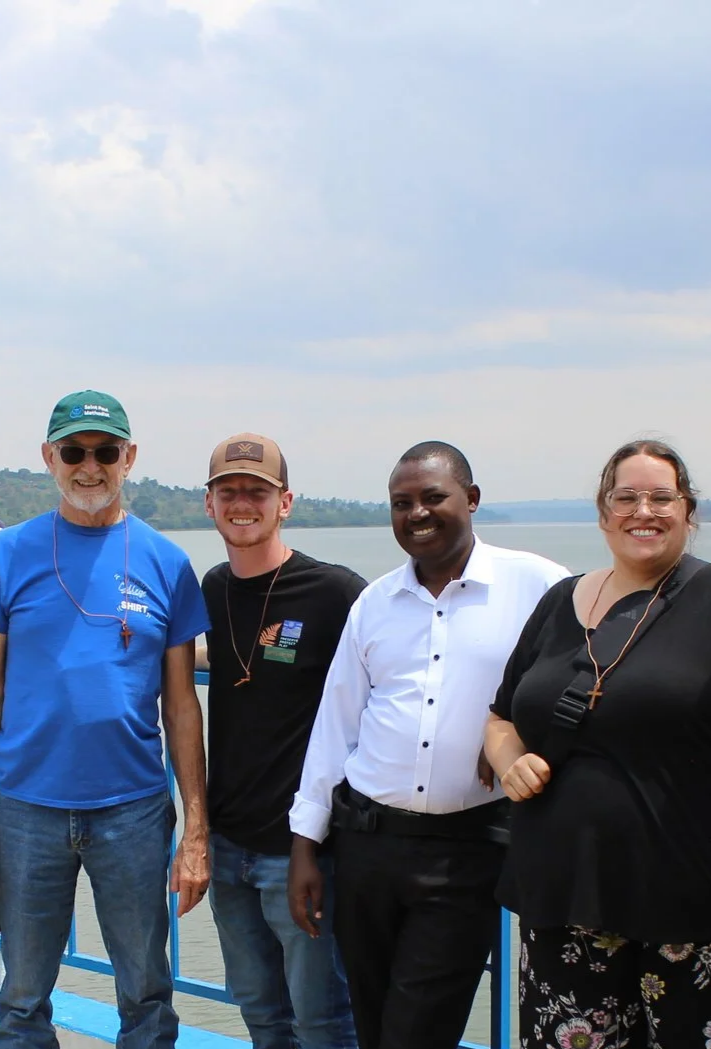 Four people standing outdoors near a body of water with a cloudy sky behind them, smiling for a photo.