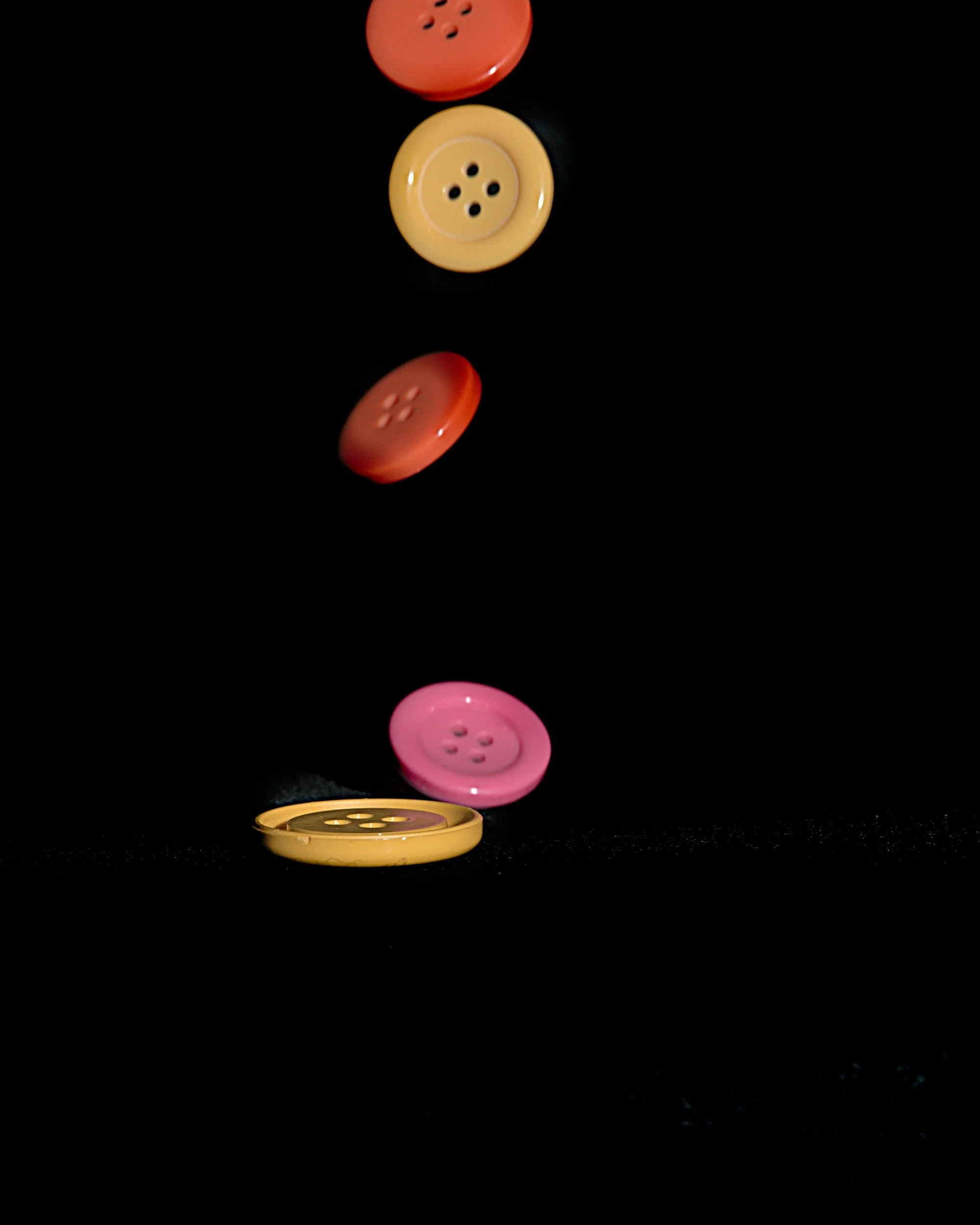 Colorful buttons in red, yellow, and pink with four holes, arranged vertically against a black background, with some appear to be falling or floating.