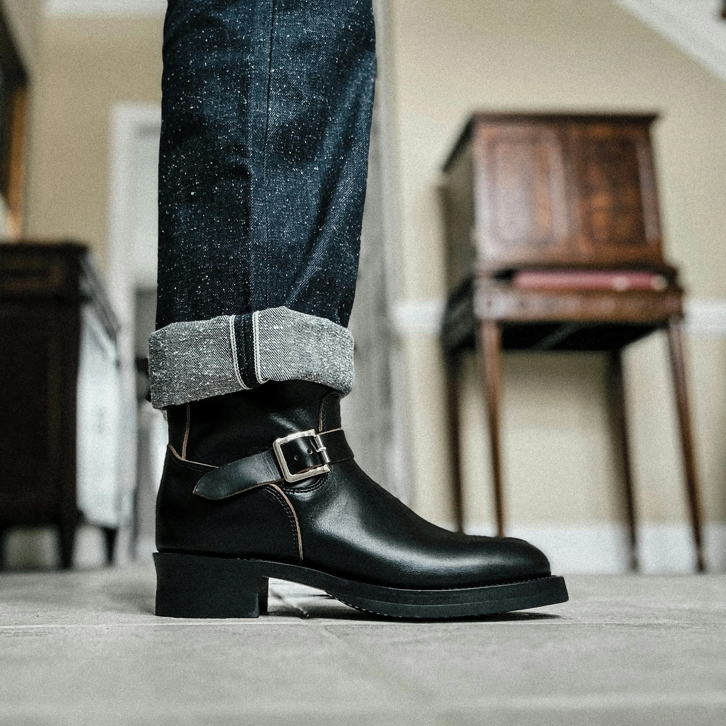 Close-up of a black leather ankle boot with a buckle, worn with rolled-up dark jeans with a splattered paint pattern, in a room with a wooden cabinet and a cabinet in the background.