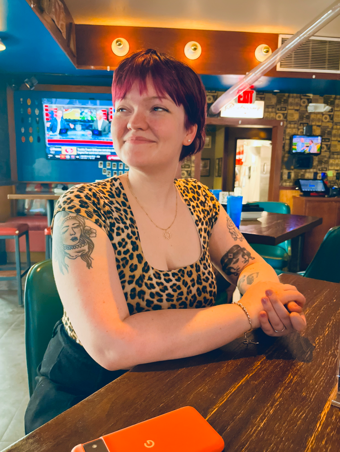A woman with short red hair and tattoos on her arms sitting at a bar or restaurant table, smiling and looking to her right. She is wearing a leopard print top and a gold necklace, with a blue drink in front of her. The background shows a TV screen, bar area, and colorful decor.