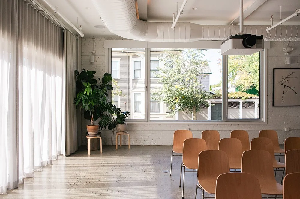 The CoLab: A bright room with large window, wooden chairs arranged facing forward, potted plants, and natural light.
