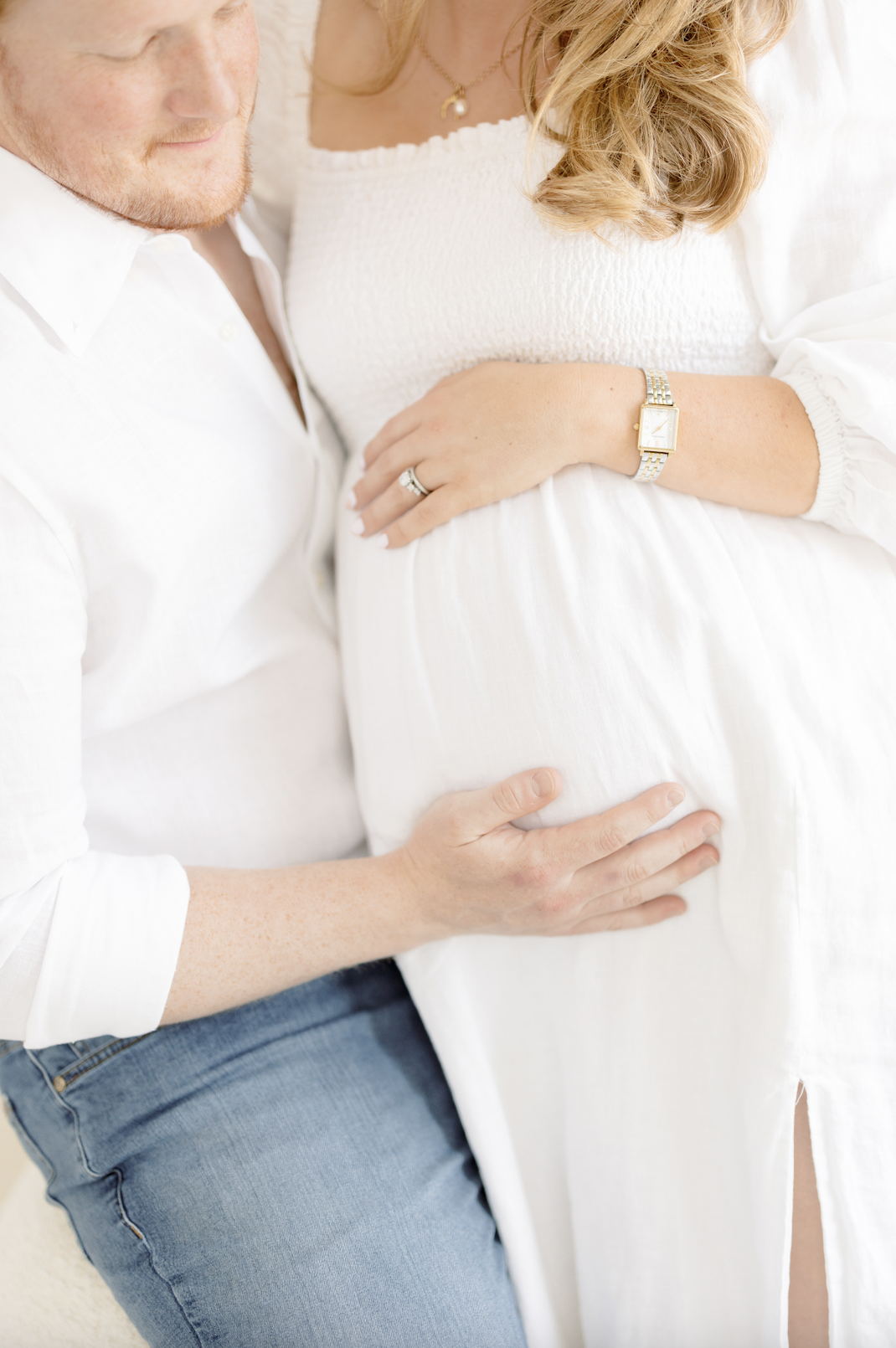 A pregnant woman with long, curly blond hair wears a white dress and a gold watch. A man, wearing a white shirt and jeans, gently holds her pregnant belly with his hand. They are close together, and her hand shows an engagement ring.