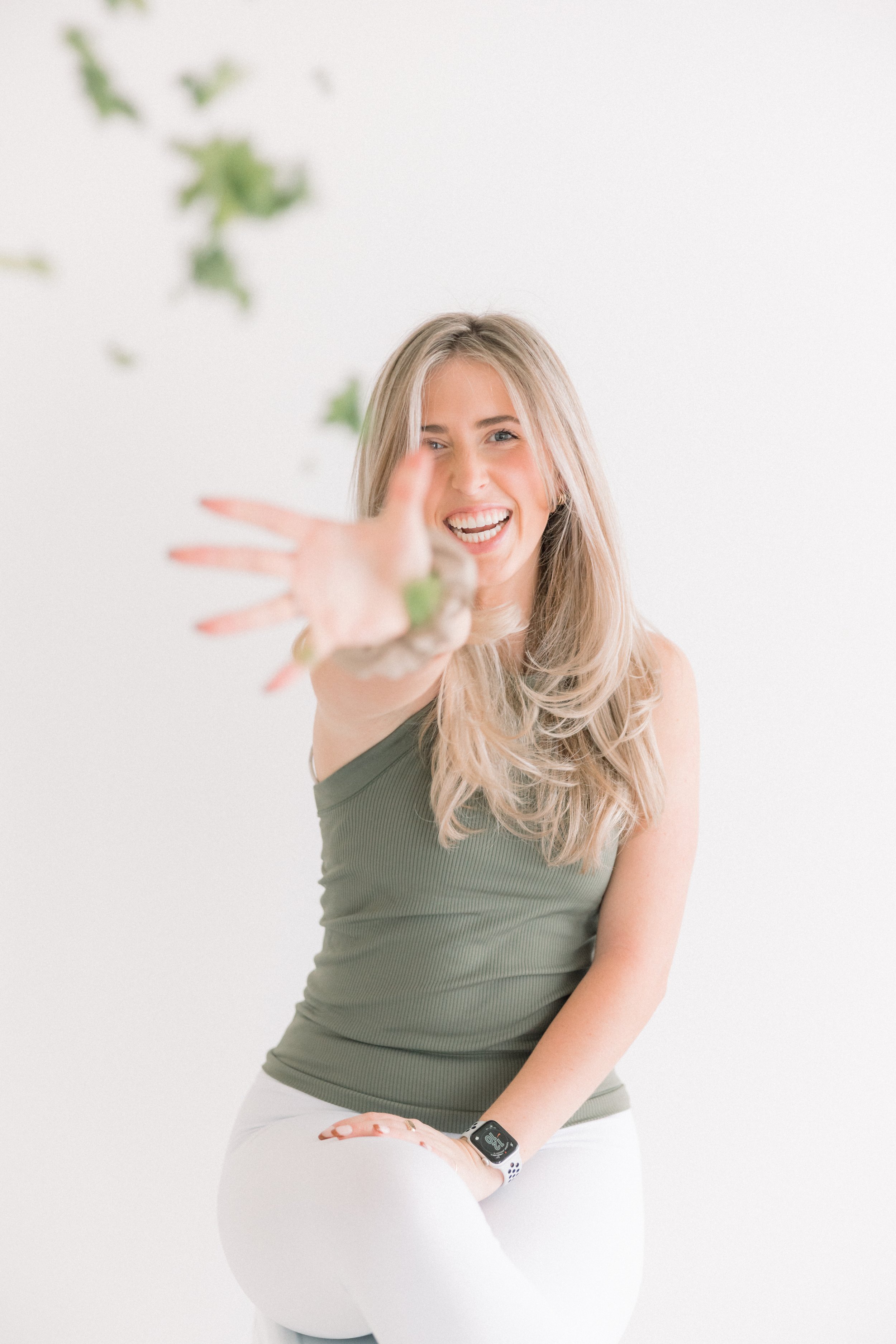 A woman with long blonde hair smiling and reaching towards the camera, wearing a green tank top and white pants, set against a plain white background with some green leaves.