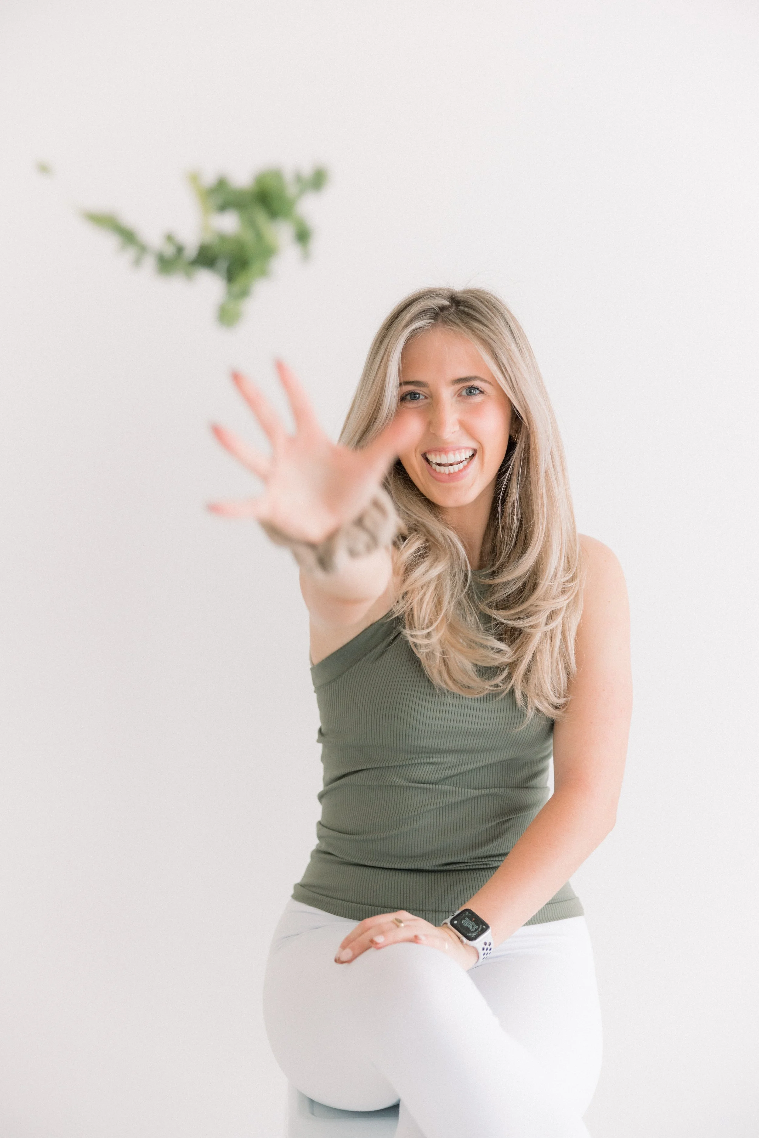 Young woman smiling and reaching towards the camera, sitting on a white surface, with a white background and blurred green leaves in the foreground.