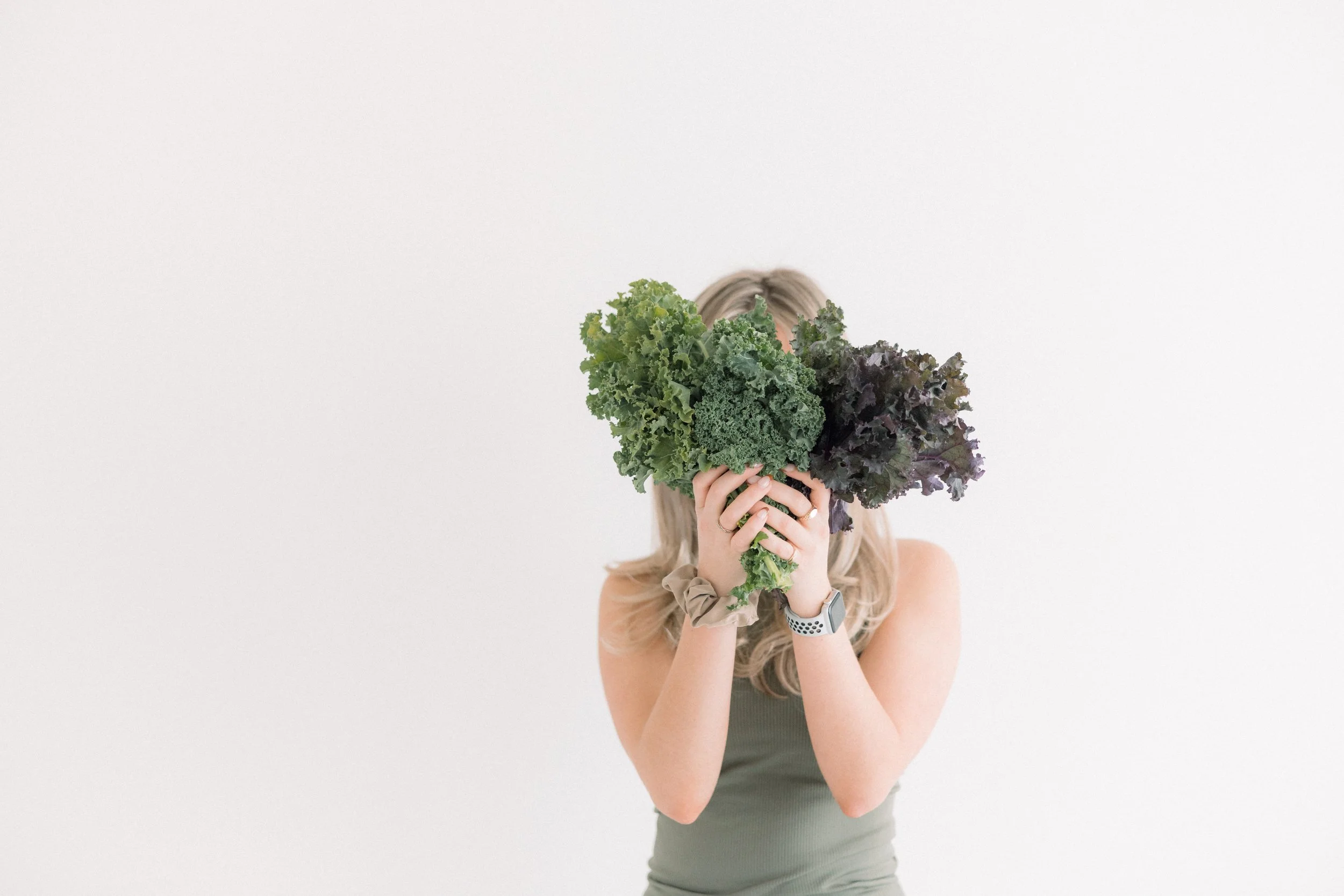 A woman with long blonde hair holding a head of green and purple lettuce in front of her face, obscuring her identity, against a plain white background.