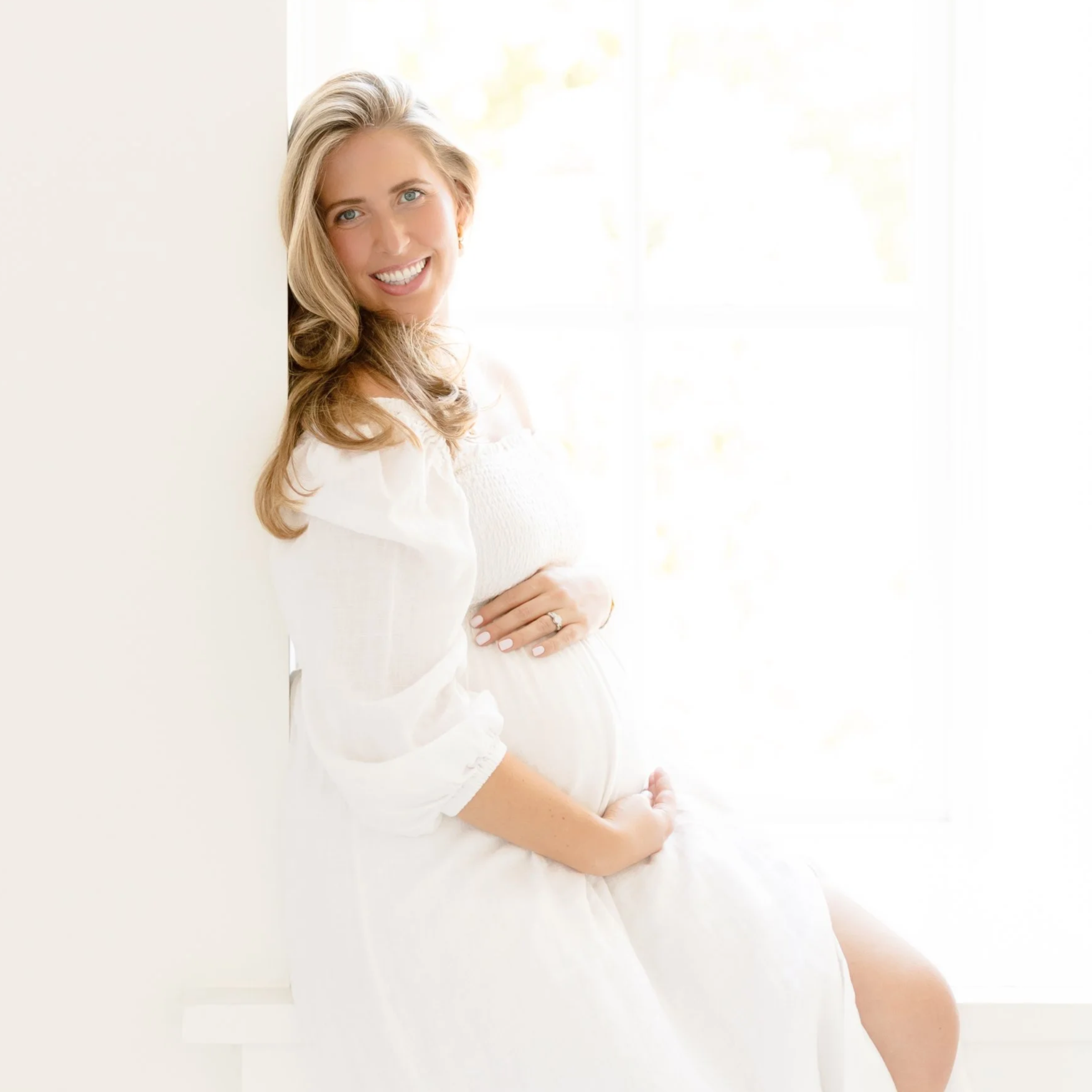 A pregnant woman with long, wavy blonde hair smiling, sitting near a window in a white dress, with her hands gently resting on her belly.